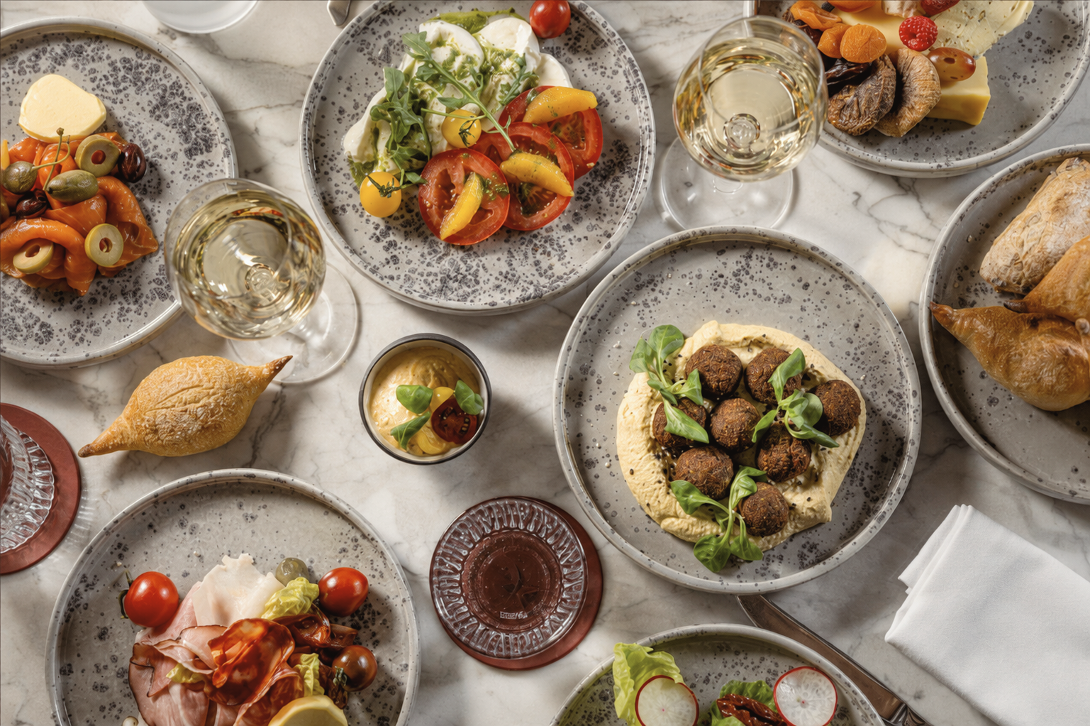 Mediterranean dining spread with hummus and falafel, fresh tomato and burrata salad, olives, artisan bread, charcuterie, and white wine.