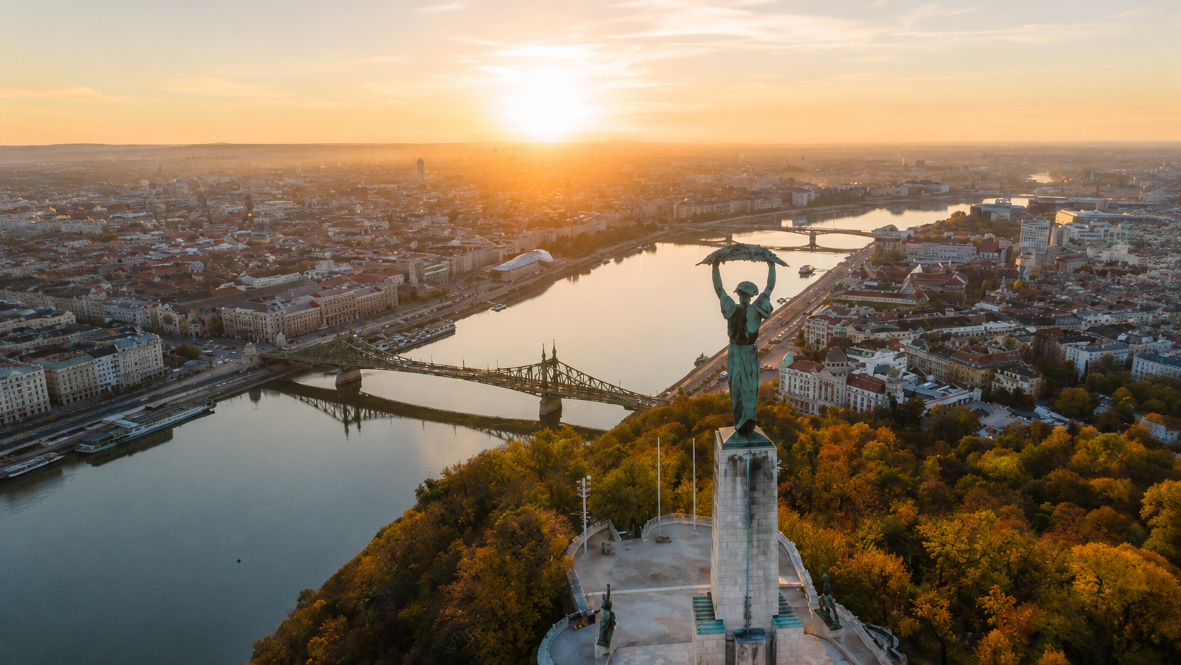 Aerial sunset view over Budapest, Hungary, from Gellért Hill, featuring the Liberty Statue, Danube River, Liberty Bridge, and historic city skyline.