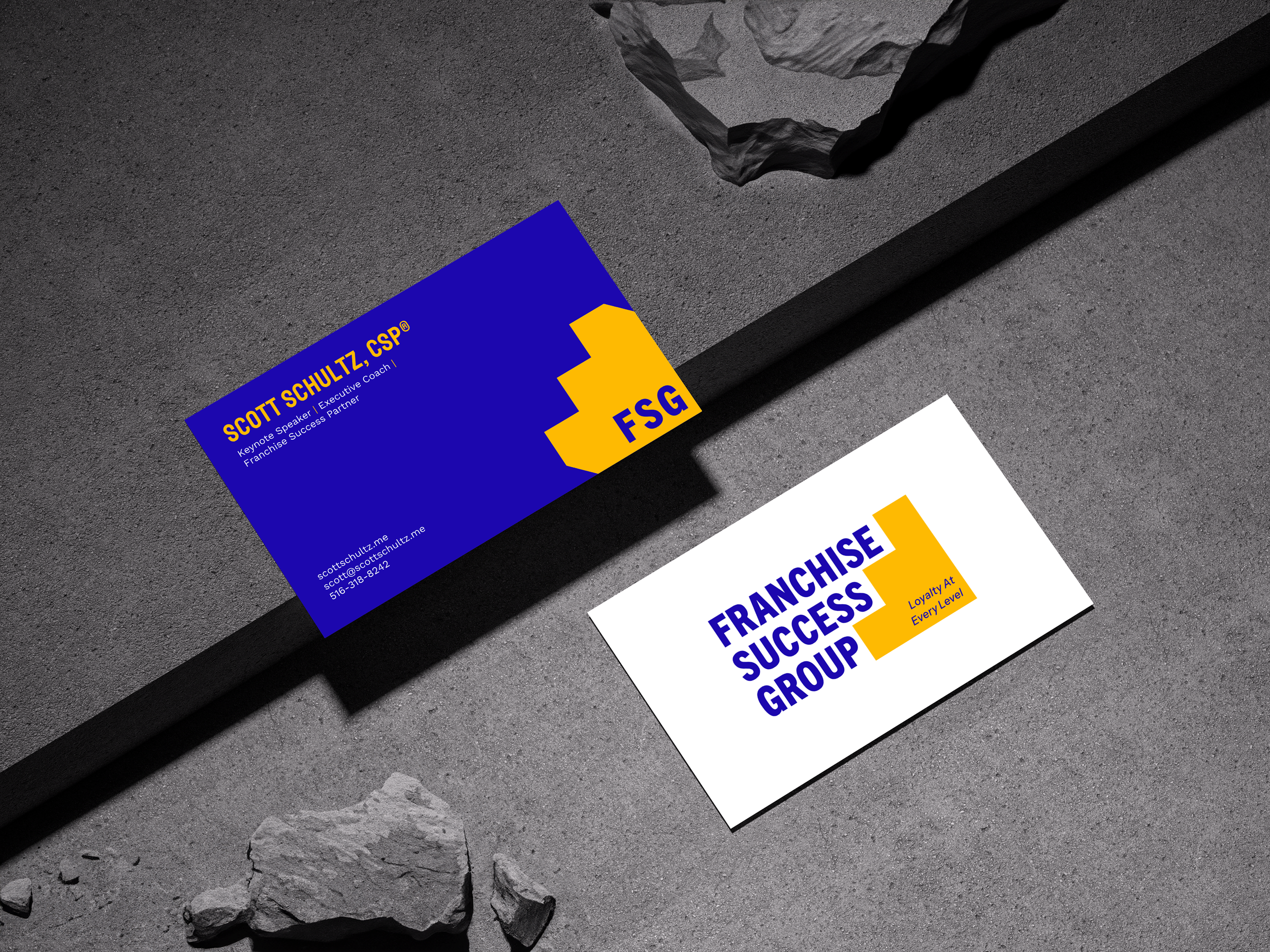 Two business cards for Franchise Success Group are placed on a gray textured surface, with rocks around them. One card is blue with orange and yellow text, and the other is white with blue and orange text.