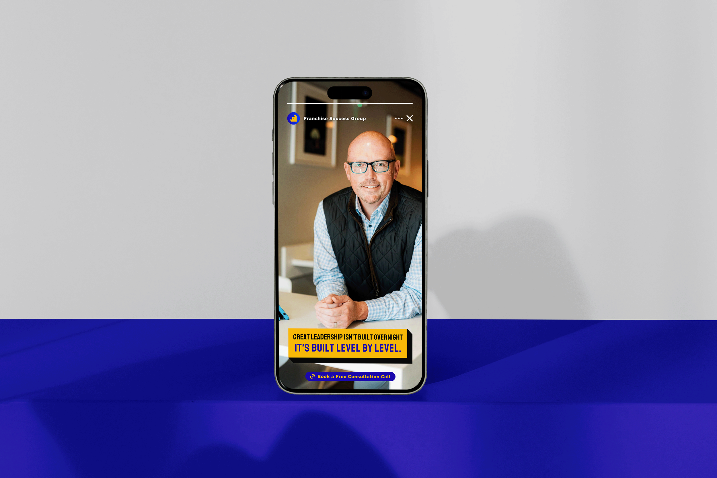Smartphone displaying a man with glasses and a blue and white checkered shirt, sitting at a table against a blurred background with framed pictures, with a yellow and black banner that says 'Great leadership isn't built overnight. It's built level by