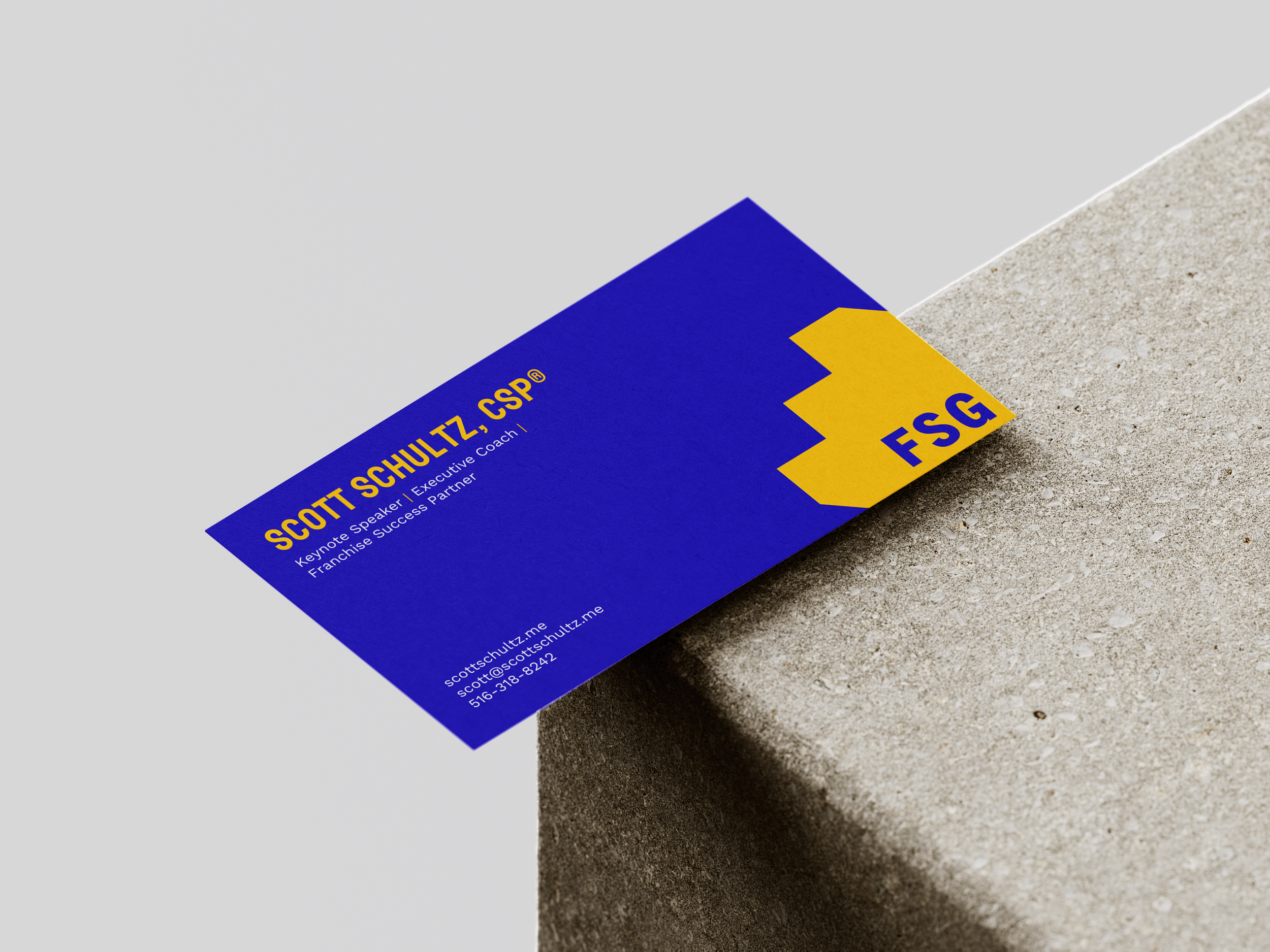 A business card for Scott Schultz CSP rests partially on a concrete surface. The card is primarily blue with yellow accents and contains contact information, including an email address and phone number.