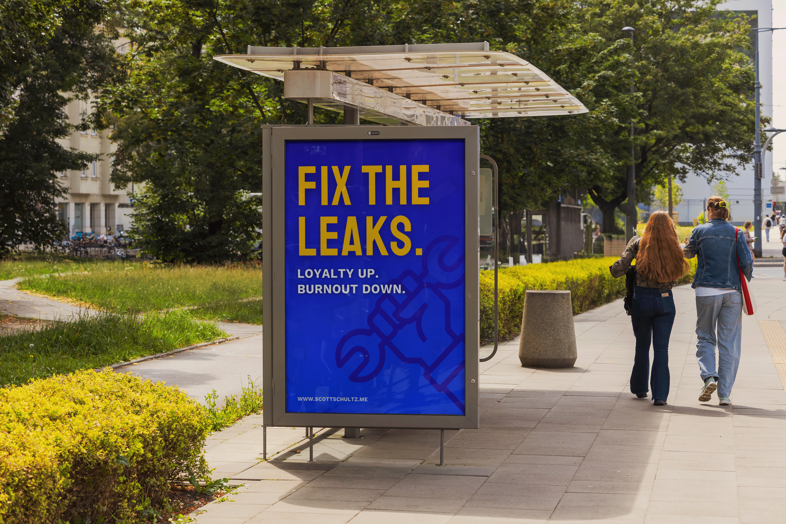 Bus stop advertisement board with a blue background displaying the text "Fix the Leaks. Loyalty Up. Burnout Down." and a graphic of a wrench and screwdriver. Two women walk on the sidewalk near the bus stop in a city environment with trees and buildi