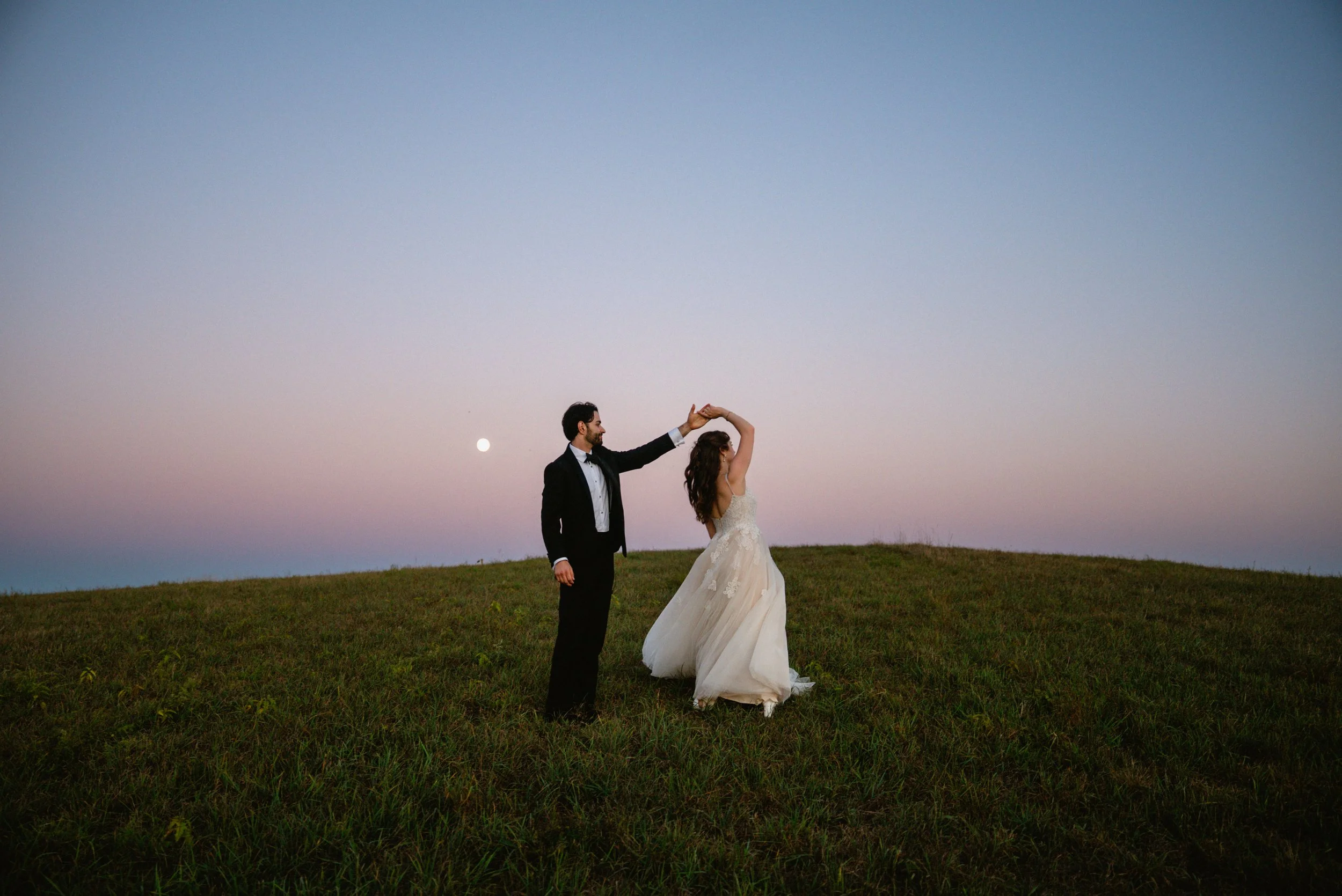 A bride and groom dancing on a grassy hill at dusk with the moon visible in the sky.