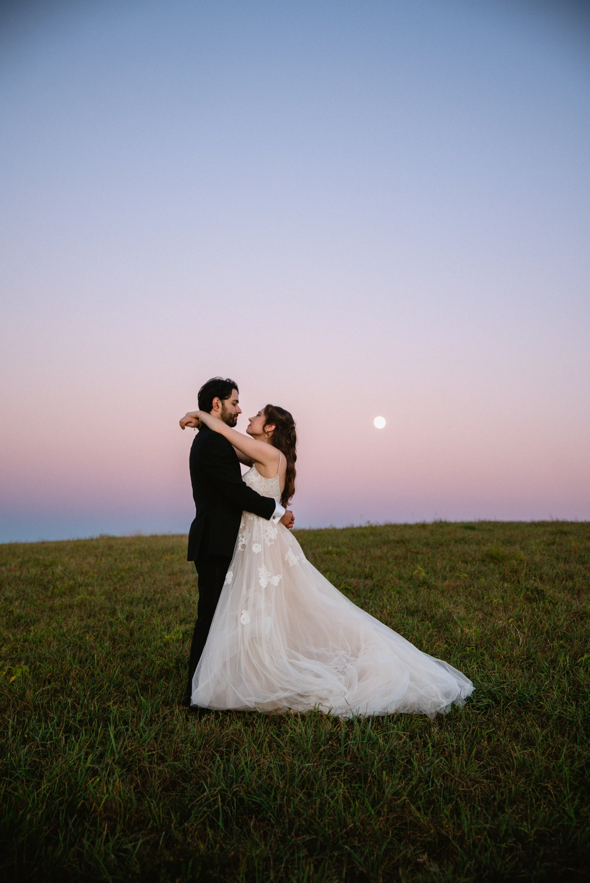 A bride and groom dancing outdoors on a grassy field at sunset, with the moon visible in the sky and a pastel-colored horizon.