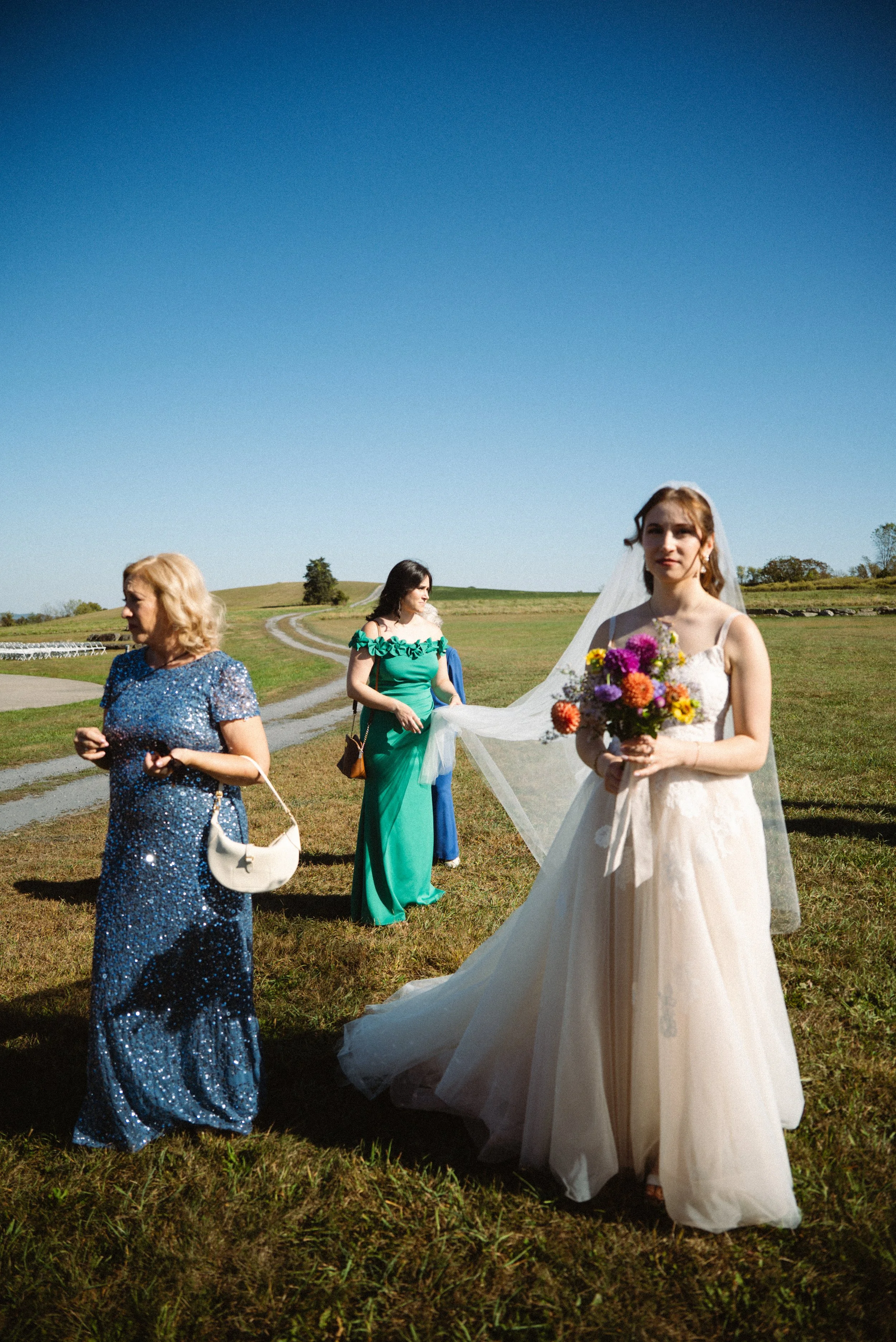 A bride in a white wedding dress holding a bouquet of colorful flowers outdoors on a sunny day, with three women in dresses nearby, walking on a grassy area with a clear blue sky in the background.