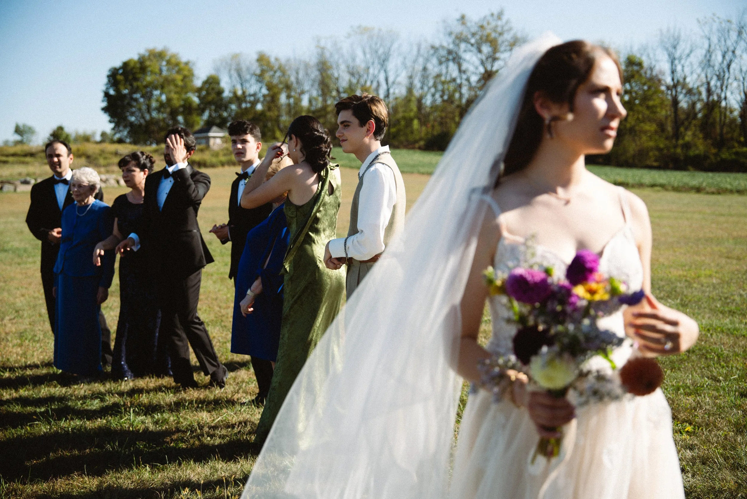 A bride in a white wedding dress with a veil holding a bouquet of purple, yellow, and white flowers, standing in the foreground. Behind her, a group of people in formal attire, mostly men in tuxedos and women in dresses, are standing and talking in a