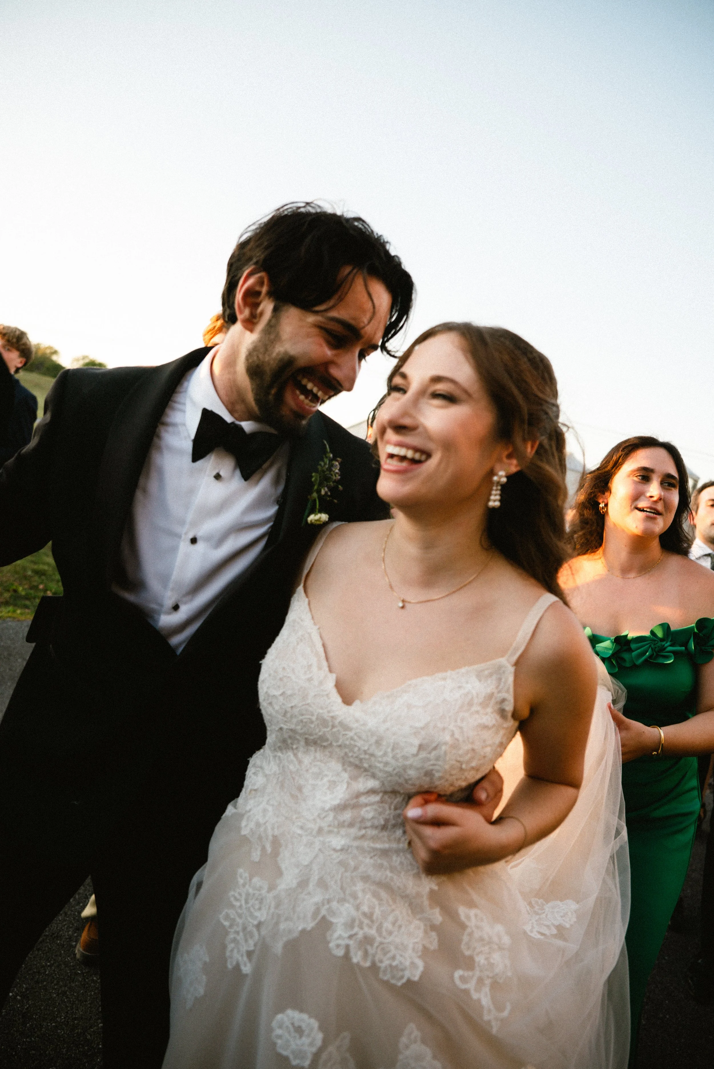 Happy bride and groom laughing together at their wedding reception, with wedding guests around them outdoors during sunset.