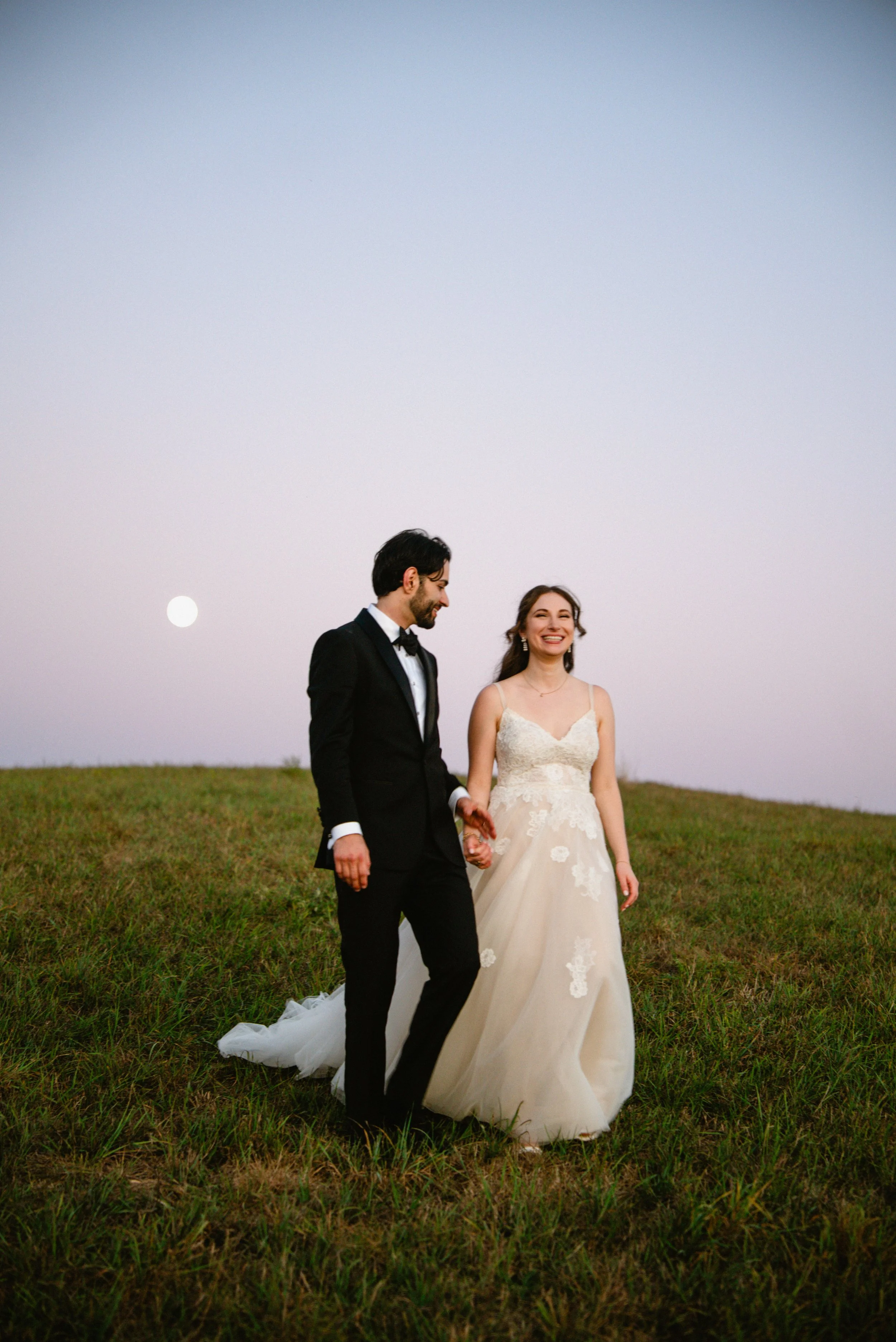 A newlywed couple walking hand in hand on a grassy hill at sunset or dusk, with the moon visible in the sky behind them. The bride wears a white wedding gown with lace details, and the groom is dressed in a black tuxedo and bow tie. Both are smiling 