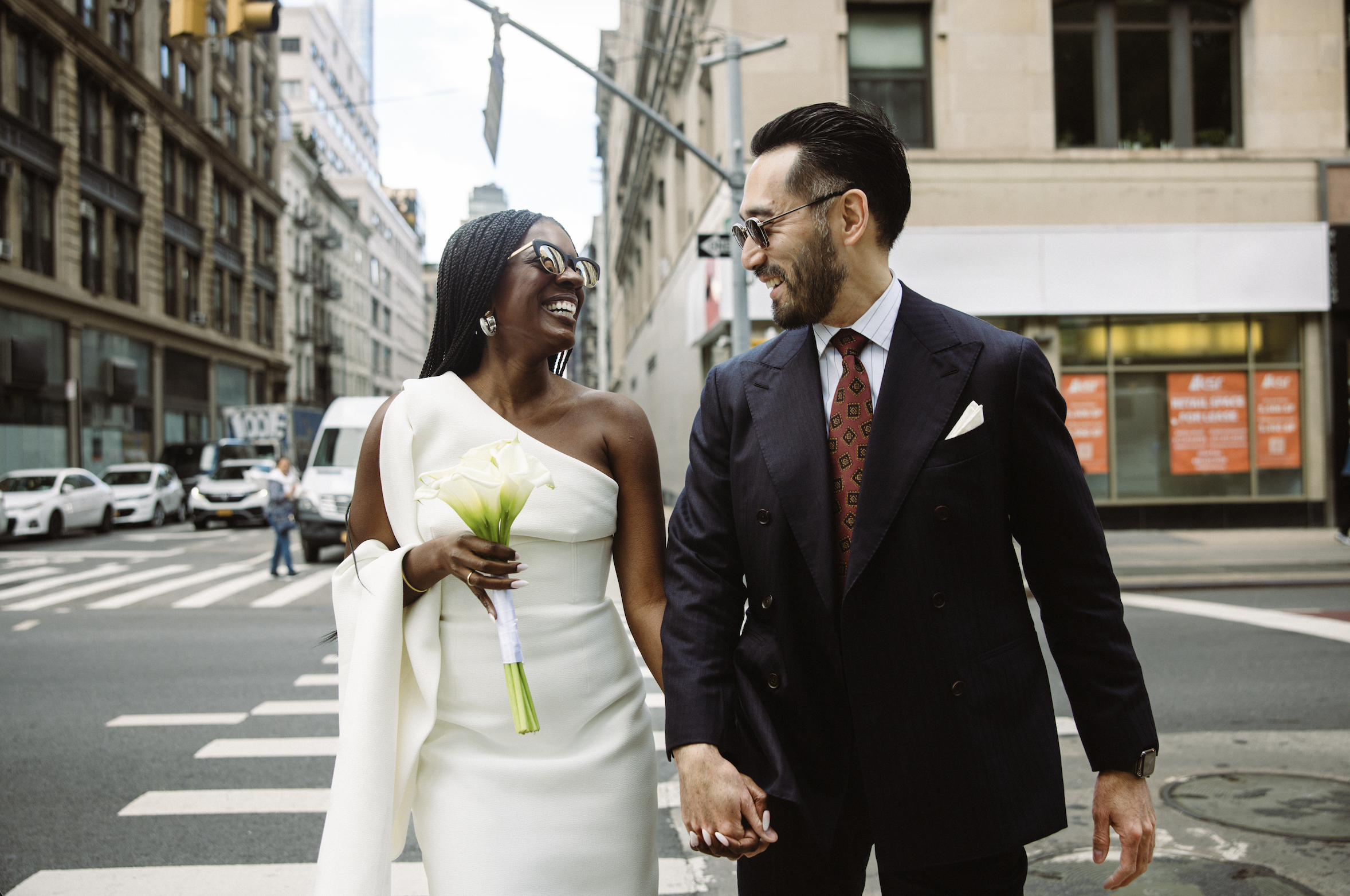 A happy couple in wedding attire holding hands on an urban street, smiling at each other, with cars and buildings in the background.