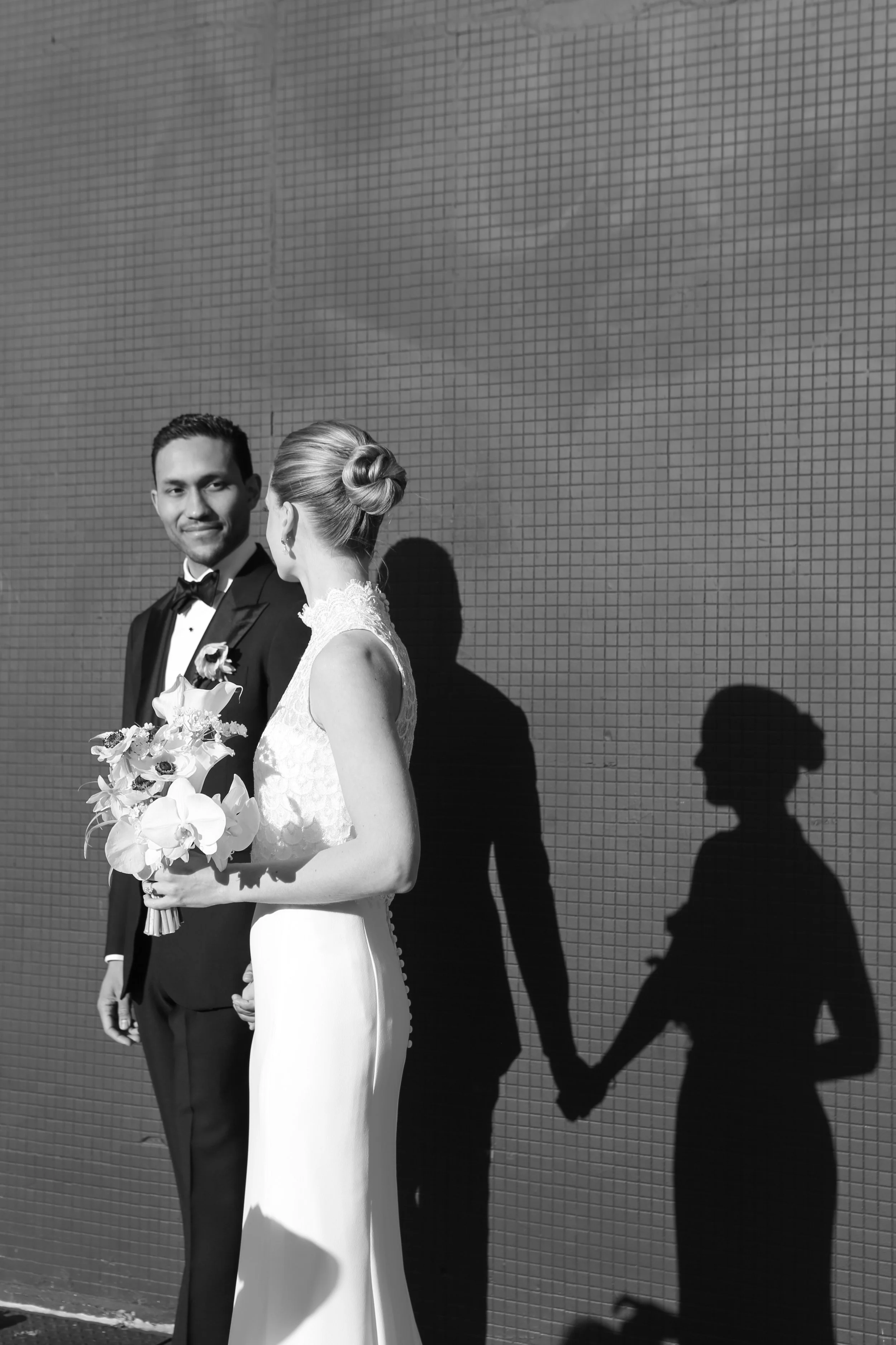 A black-and-white photo of a bride and groom holding hands against a tiled wall, with their shadows cast on the wall.