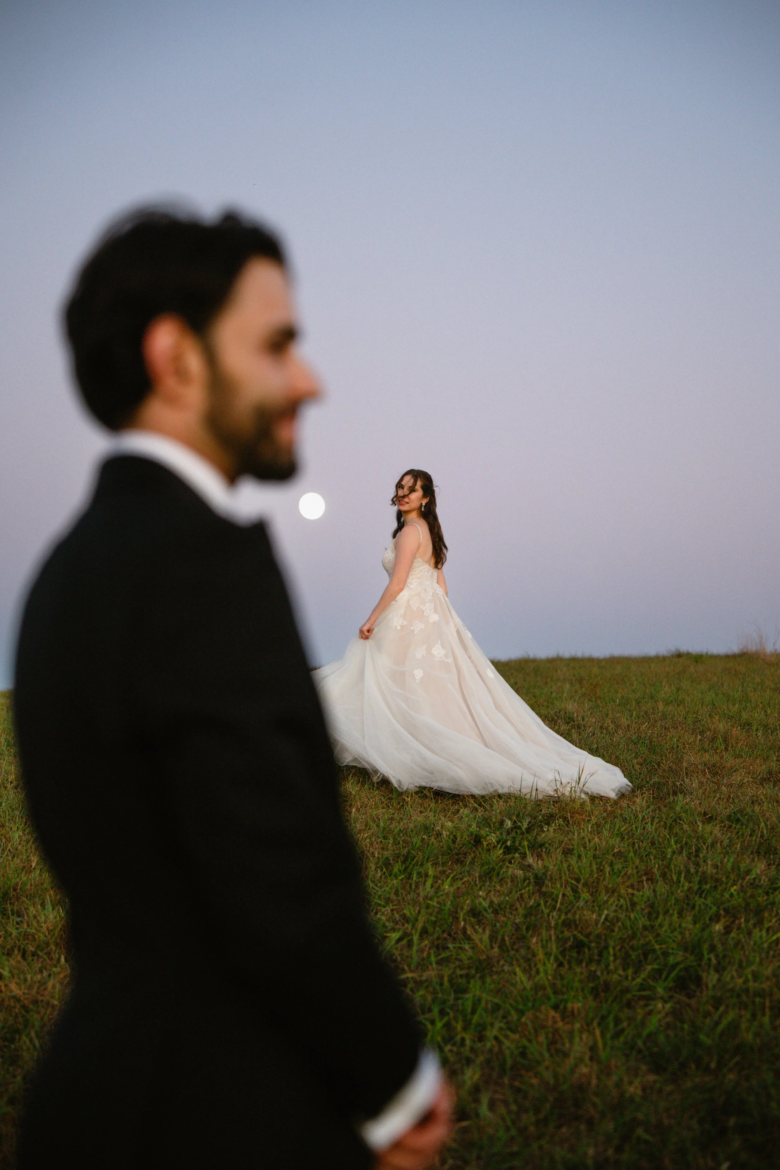 A bride in a white wedding gown standing on a grassy hill at dusk, with a bride and night sky in the background, in focus, while a groom in a black tuxedo is blurred in the foreground.