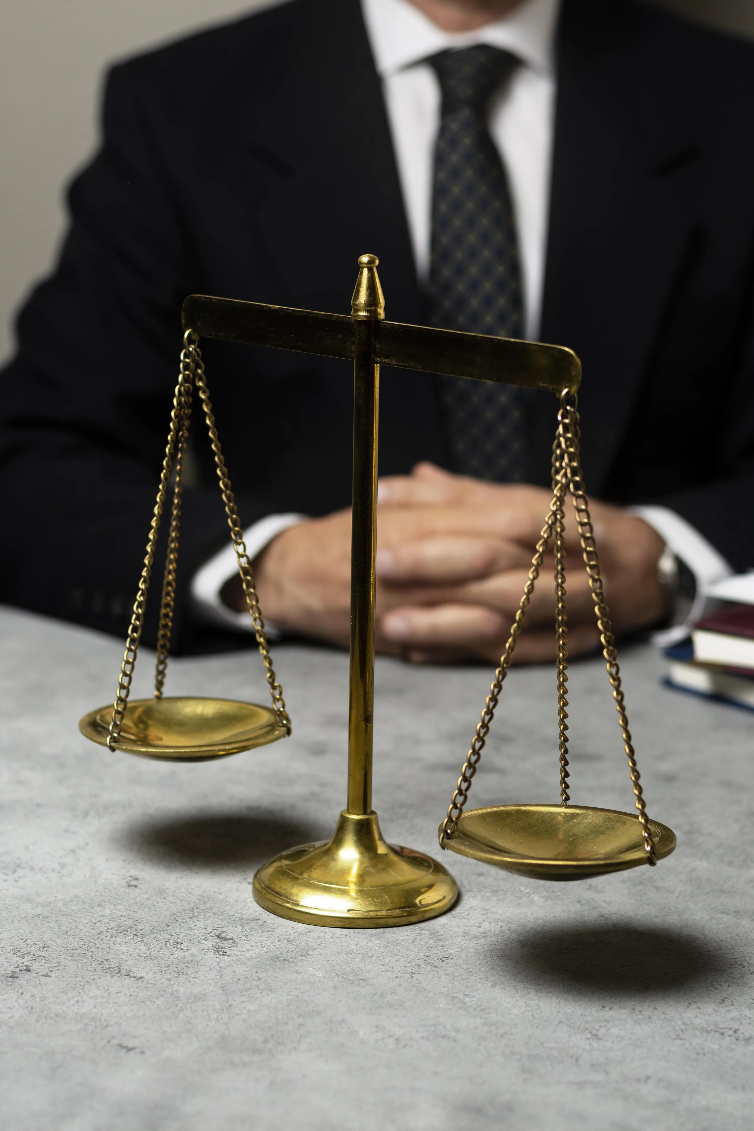 A person in a dark suit with a tie sitting behind a table with brass balance scales in the foreground.