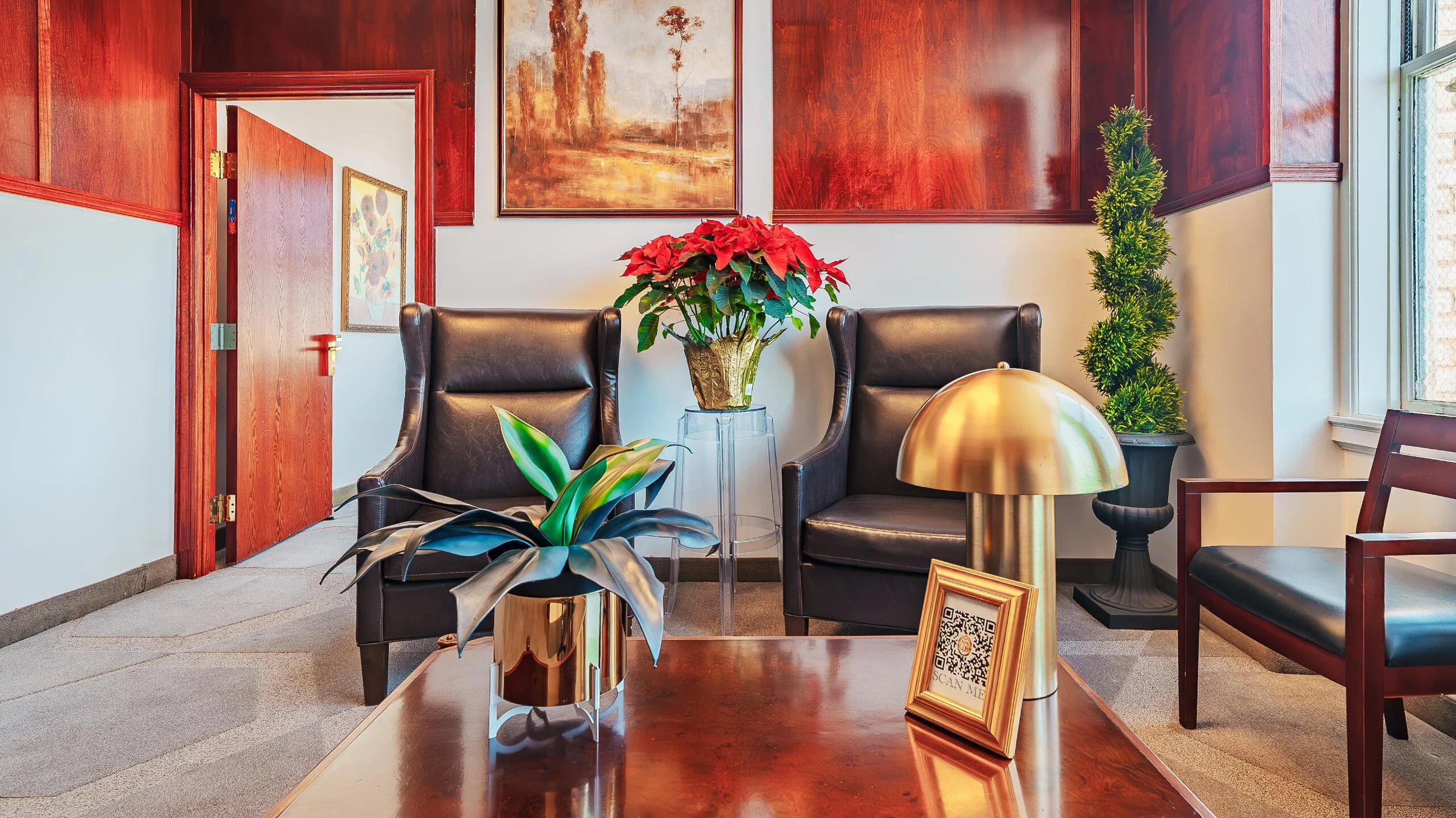 Interior of a waiting room with two black leather chairs, green indoor plants, a wooden table with a potted red poinsettia, a gold-toned table lamp, framed artwork on the wall, and a QR code sign on the table.