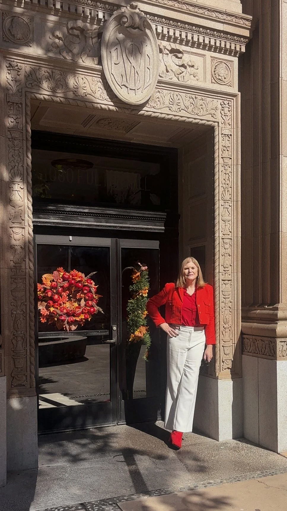 A woman wearing a red jacket, red blouse, red shoes, and white pants stands outside an ornate building with a decorative stone arch and a crest above the doorway. She is next to a black vase with fall-colored autumn foliage and a ribbon wreath decoration hanging on the glass door.