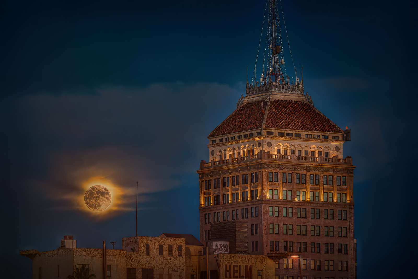 Tall building with a distinctive roof and antenna, illuminated at night, with a large moon in the cloudy night sky.