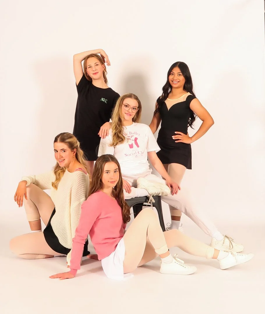 Five dancers posing together in a studio with a plain white background, some sitting on the floor, others standing, all smiling and wearing casual clothing.
