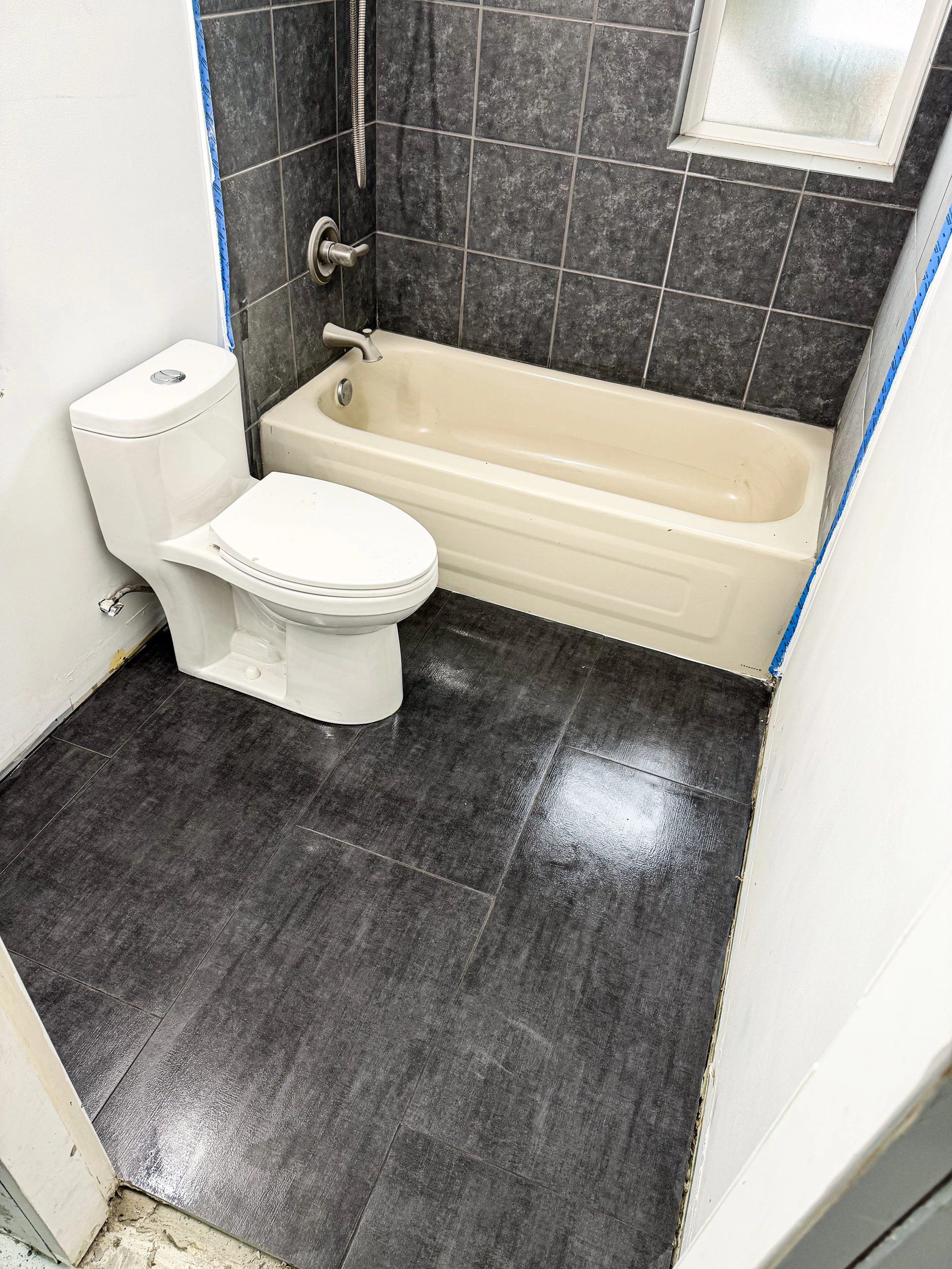 A small bathroom with dark gray tiled flooring, a white toilet, a beige bathtub, and a window with frosted glass.