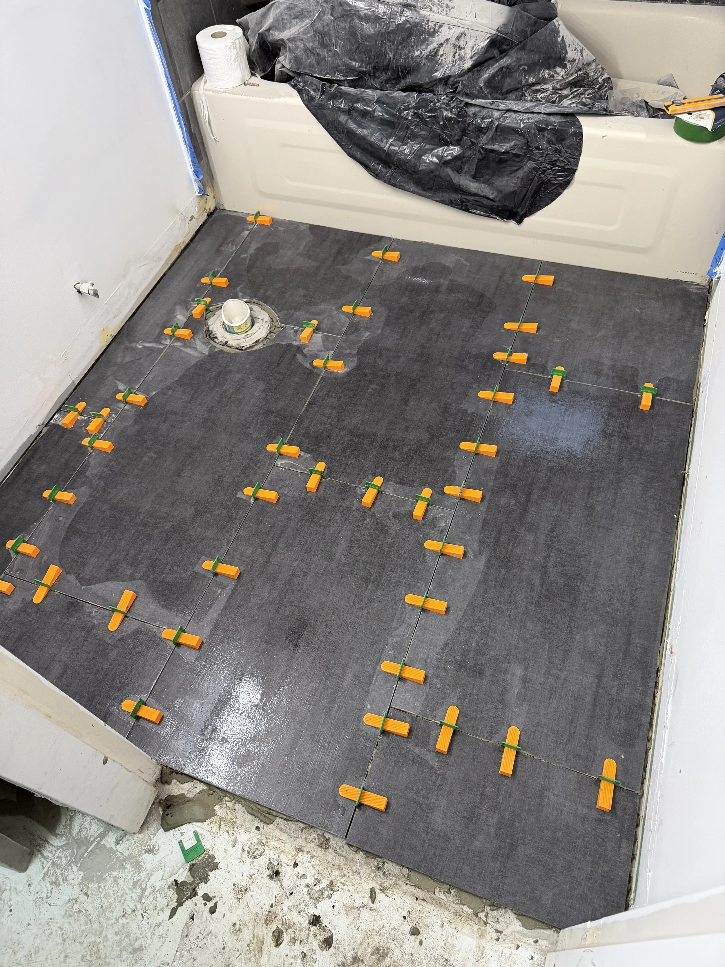 Bathroom floor with dark tiles being installed, with orange and green tile spacers evenly placed between tiles, near a bathtub with a black plastic sheet and toilet paper roll on top.