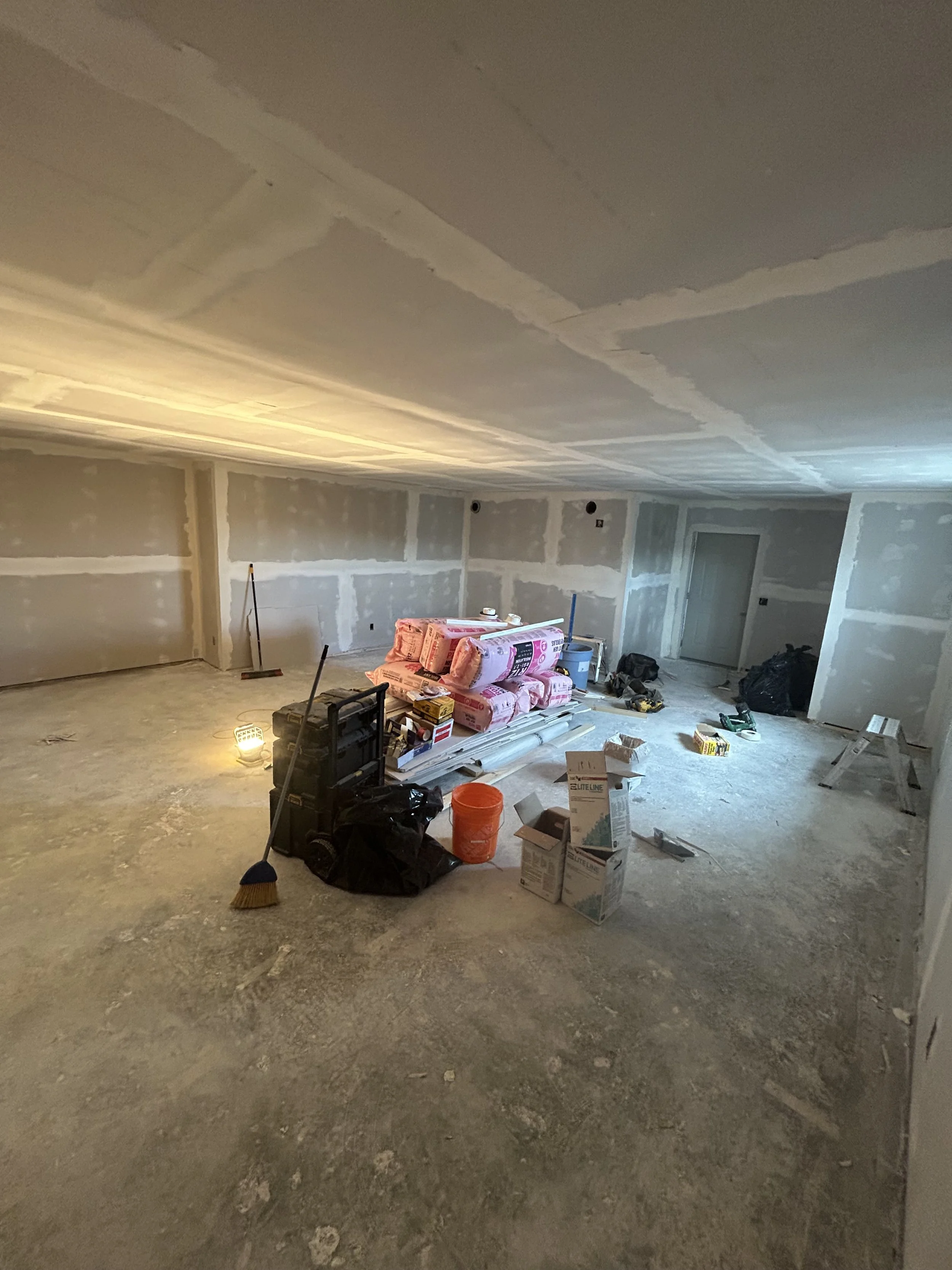 Unfinished basement room under construction or renovation with drywall walls and ceiling, construction tools, supplies, and insulation material scattered on the floor.