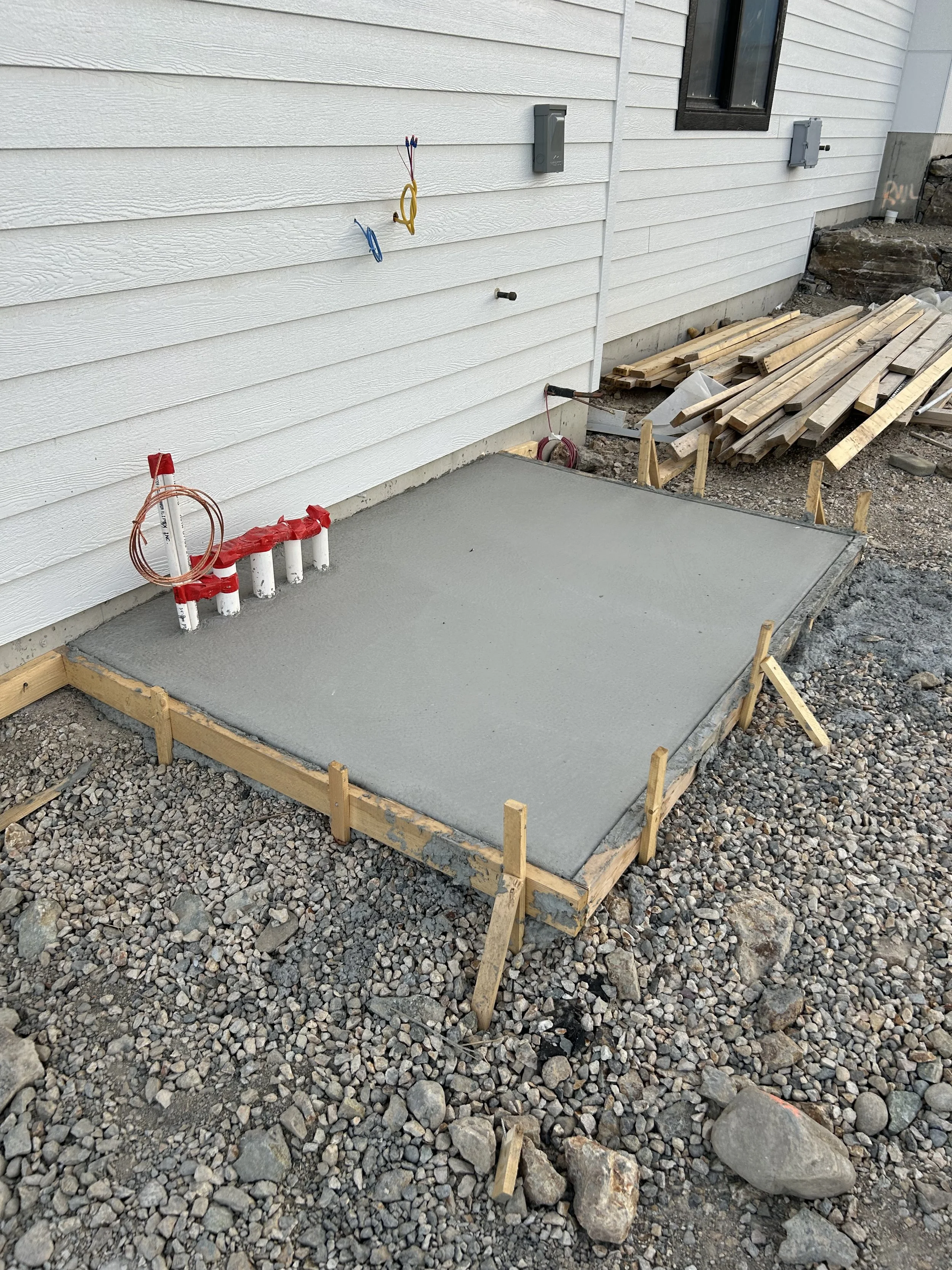 Freshly poured concrete slab foundation for a new outdoor area near a house, with wooden framing and construction materials around.