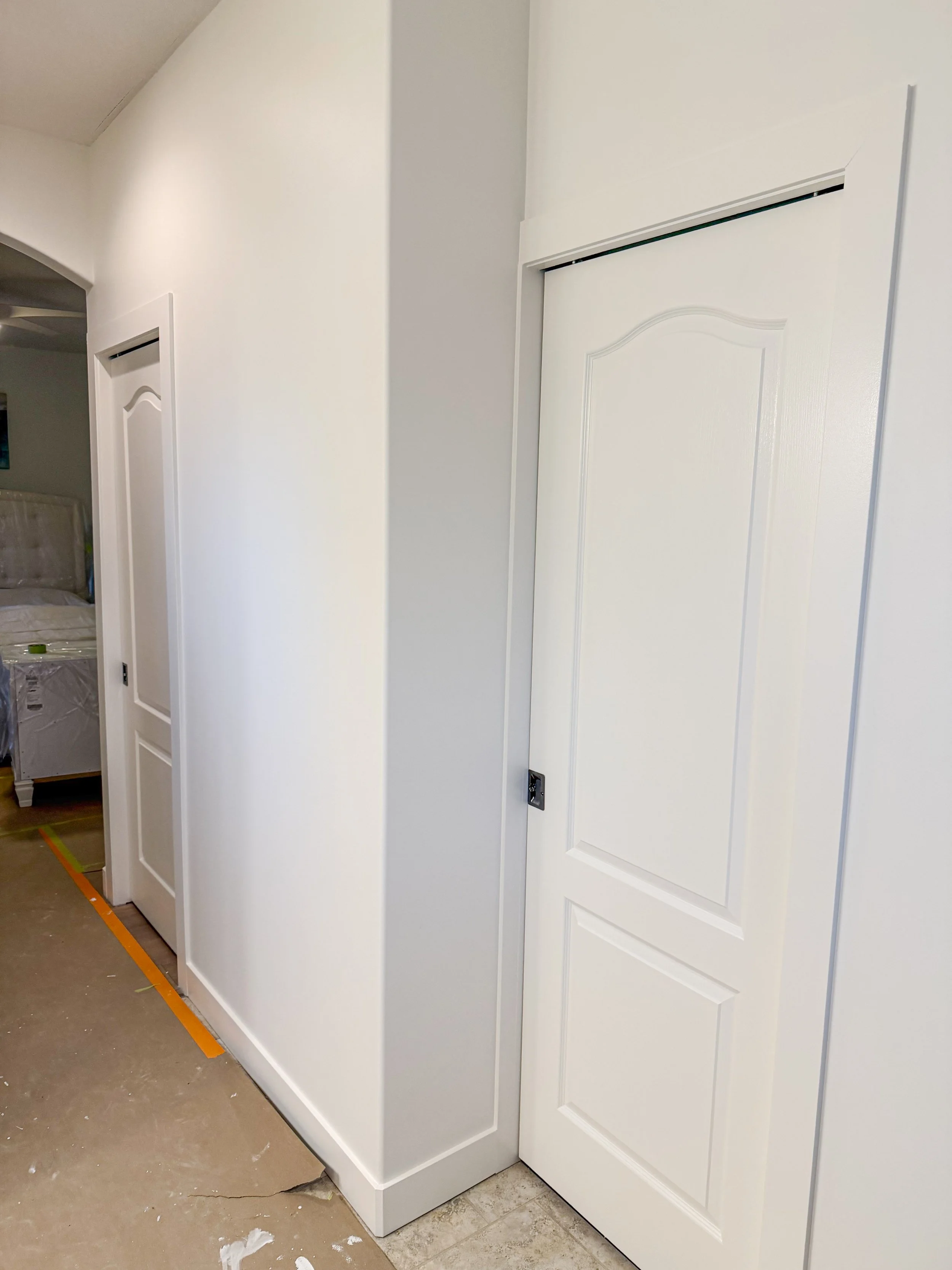 Interior view of white closet doors, with part of a doorway to another room visible on the left side, and a bed in the background.