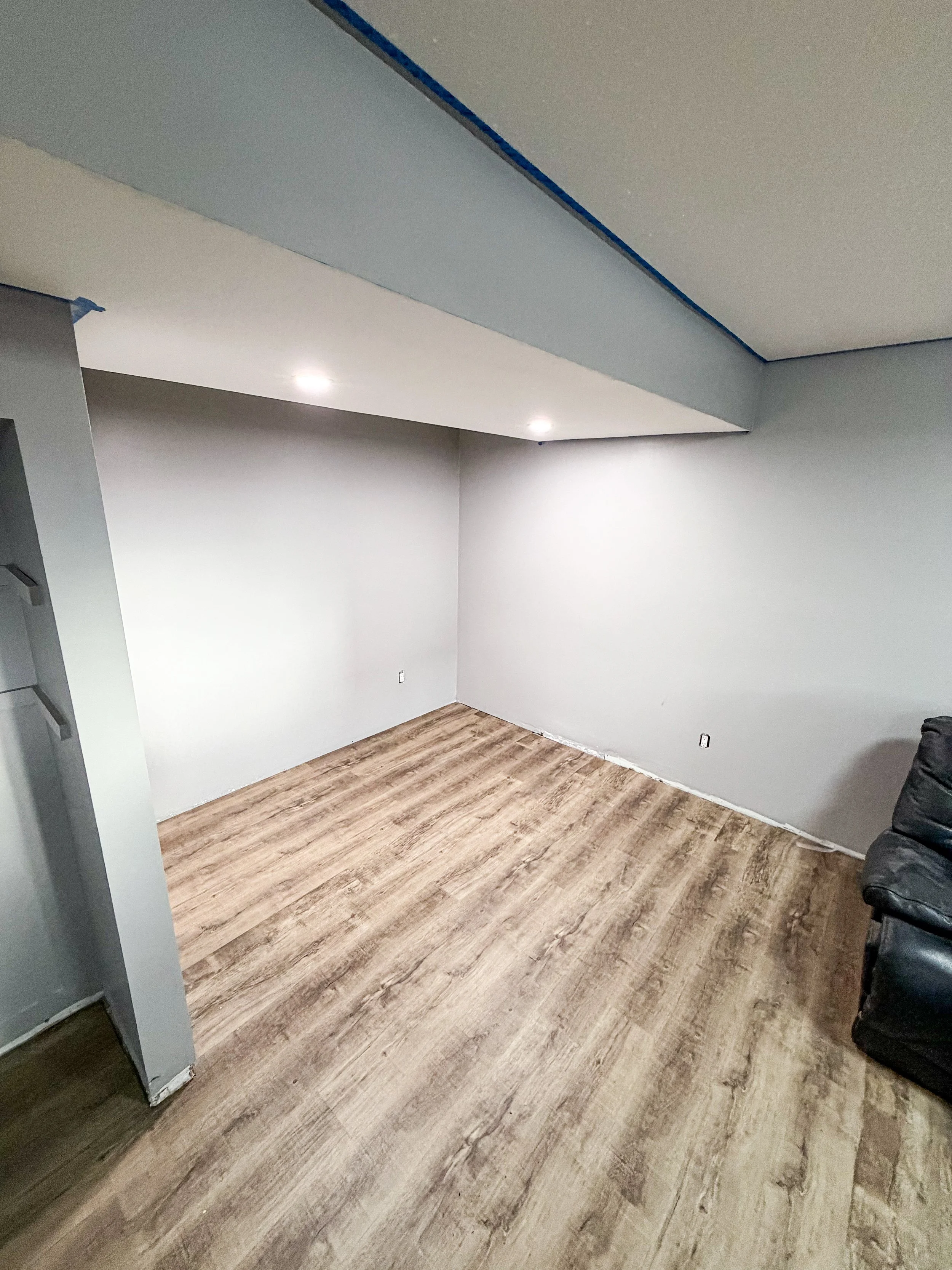 A room under renovation with gray walls, a partially finished ceiling, and wood-look flooring. There are two ceiling lights and an outlet on the wall. A black couch is partially visible on the right.