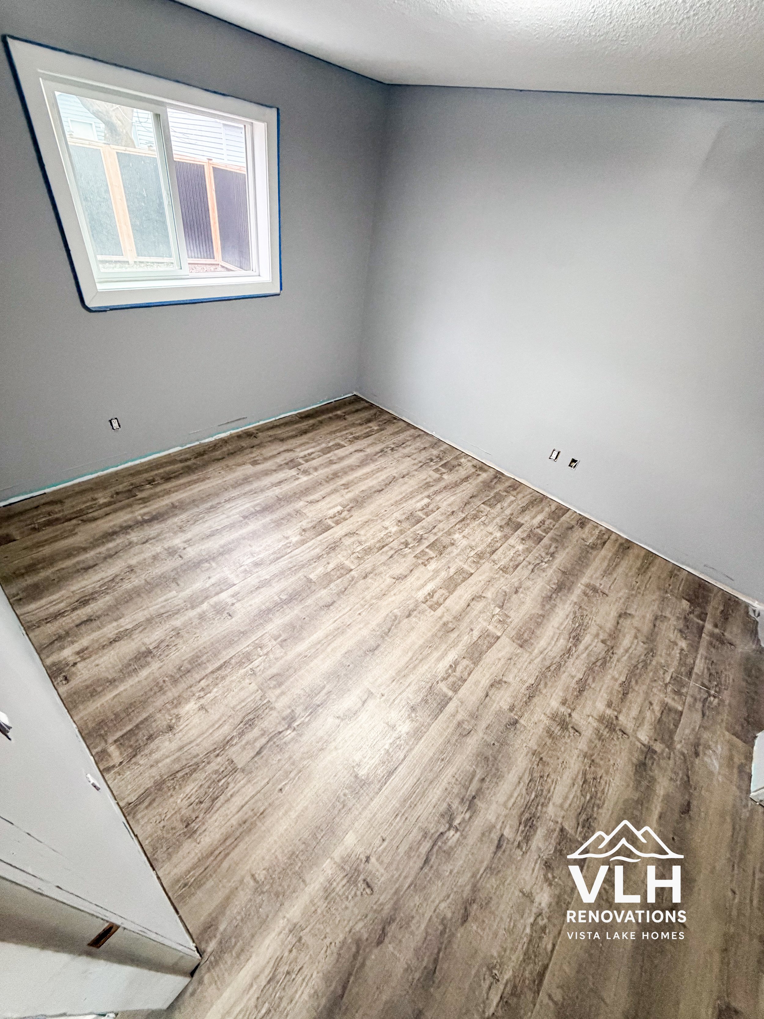 Empty room with light gray walls, a single window, and wood-style flooring during renovation.