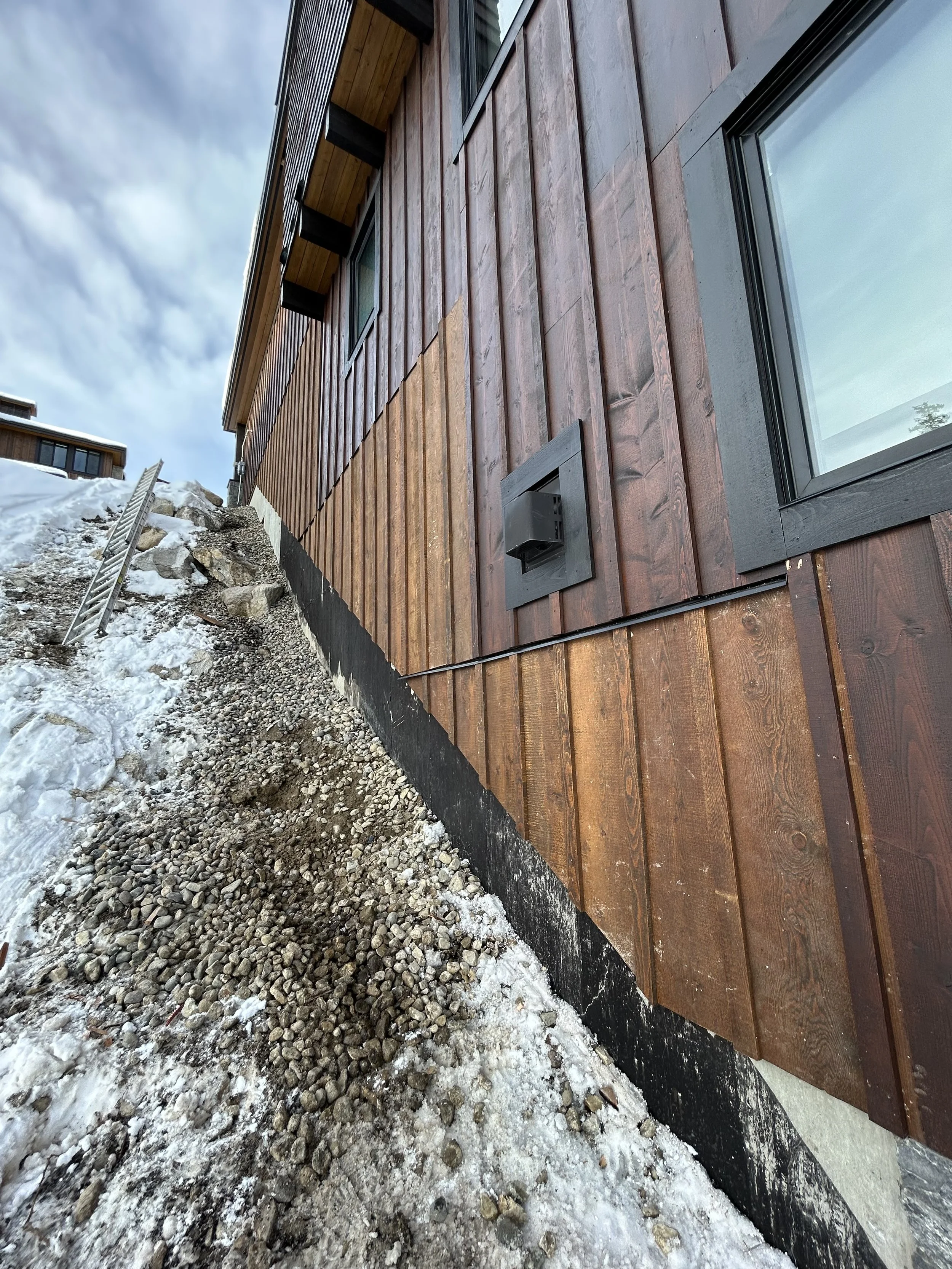 Close-up of the exterior of a building with brown wooden siding, black window frames, a black vent, and snow and rocks on the ground outside.