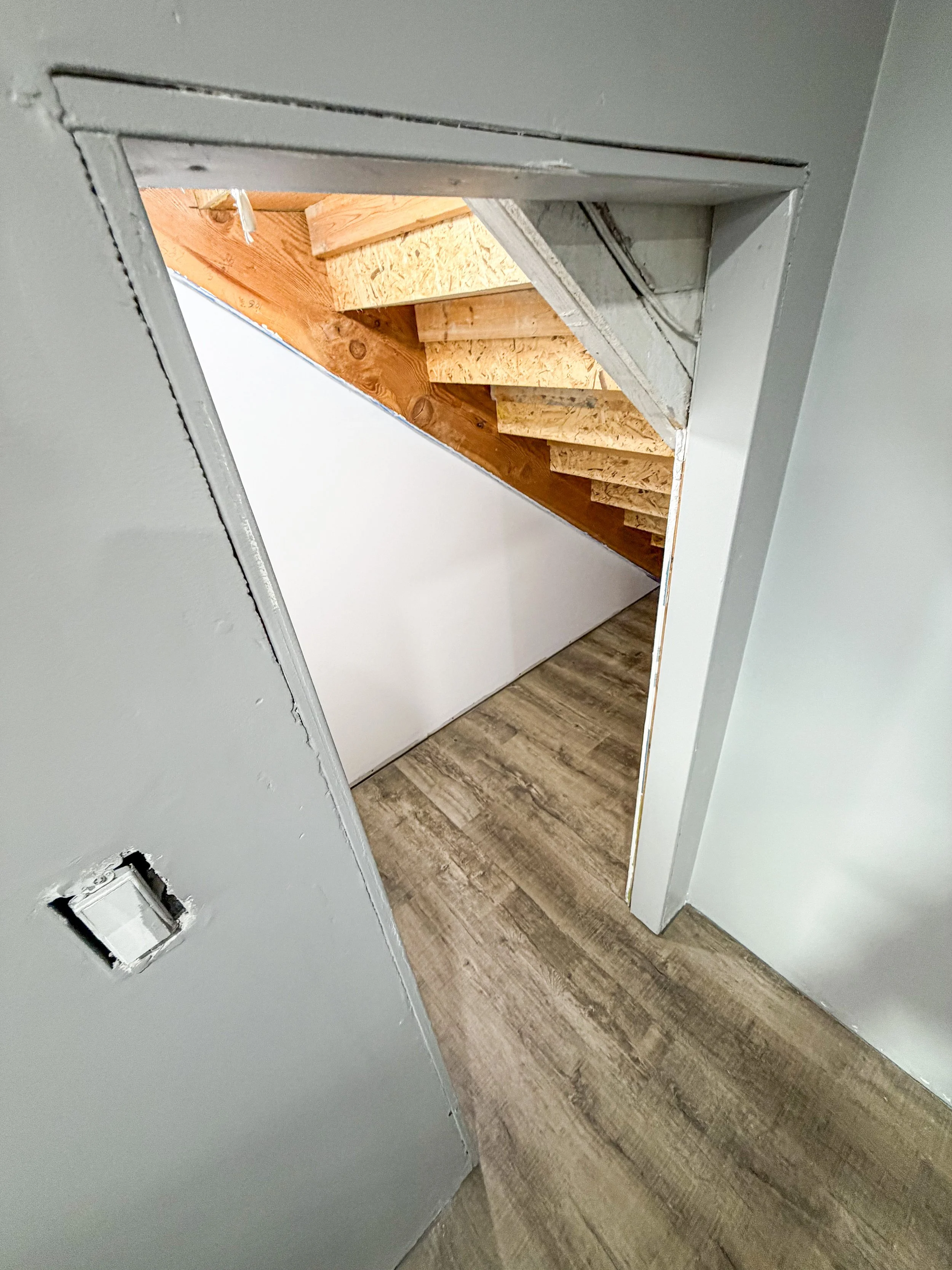 View through a doorway showing wooden staircase under construction, with exposed wooden steps, and a white wall on one side, on a hardwood floor.