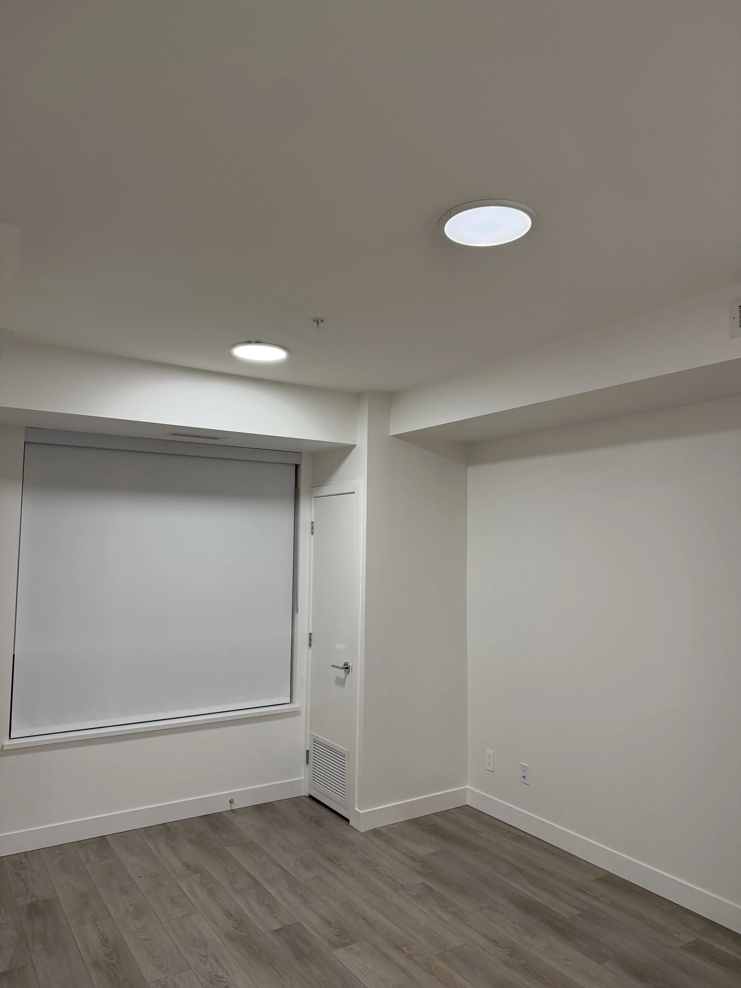 Empty white room with a large window, wooden floor, and two ceiling lights.