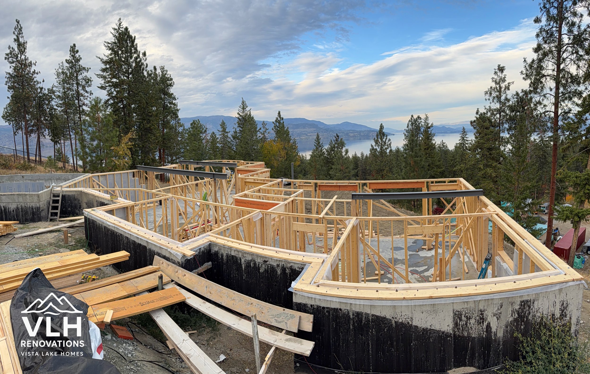 Under construction view of a house's wooden framing with a black waterproofing layer on a hillside overlooking a lake surrounded by trees and mountains.