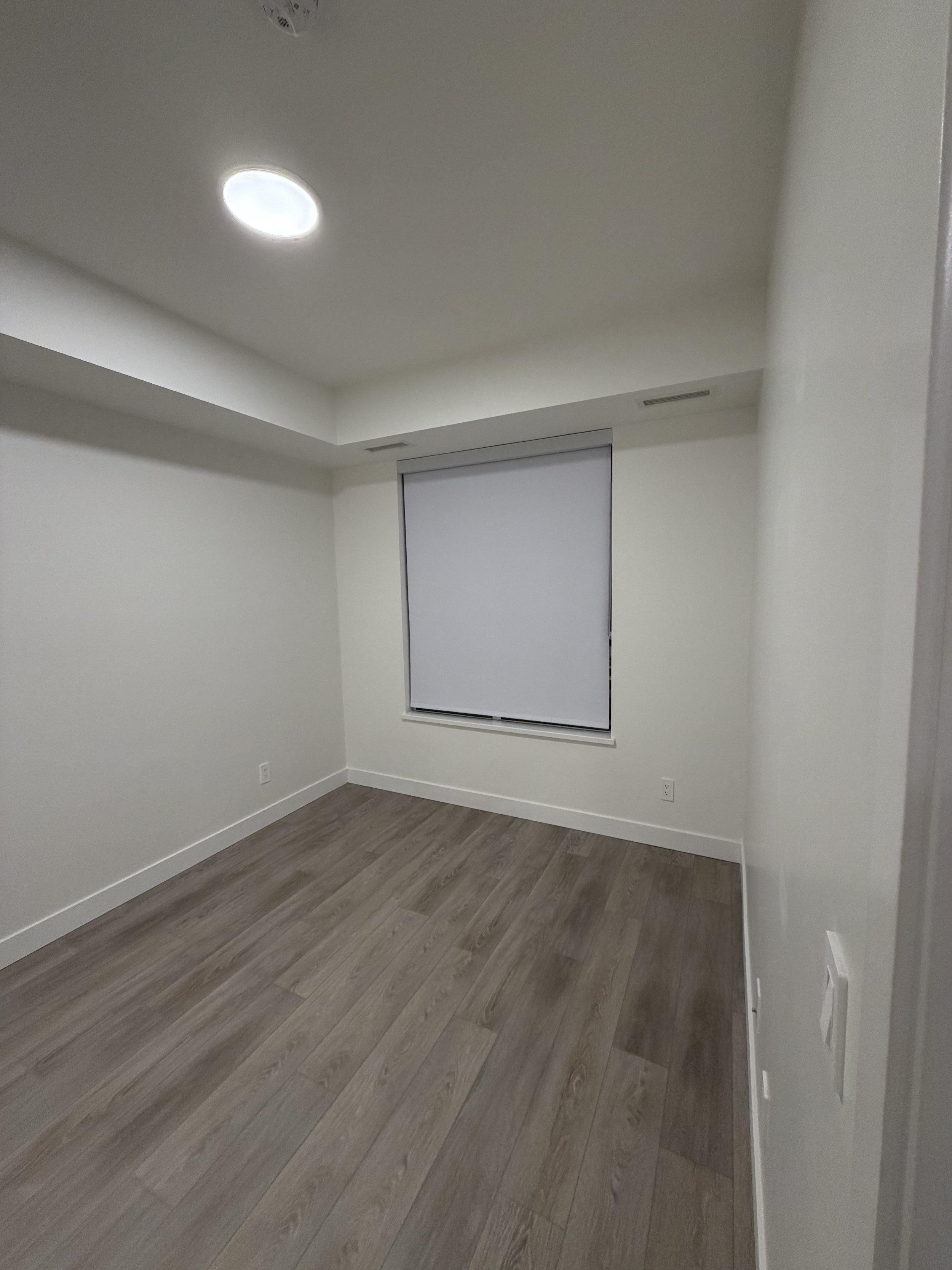 Empty room with white walls, a window with closed blinds, a ceiling light, and wood flooring.