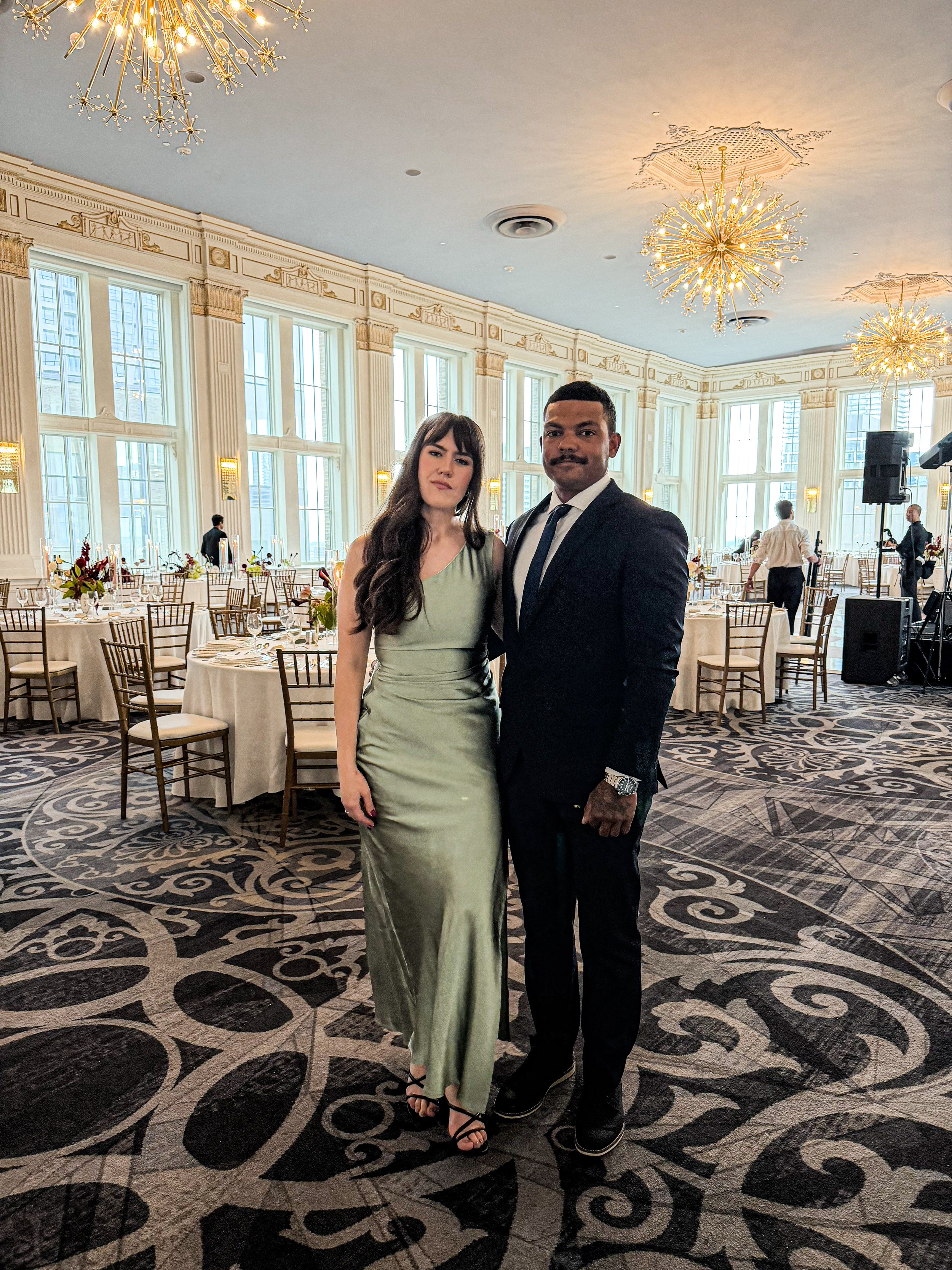 A woman in a satin green gown and a man in a black suit pose together in a decorated banquet hall with large windows, ornate chandeliers, and round tables with floral centerpieces.