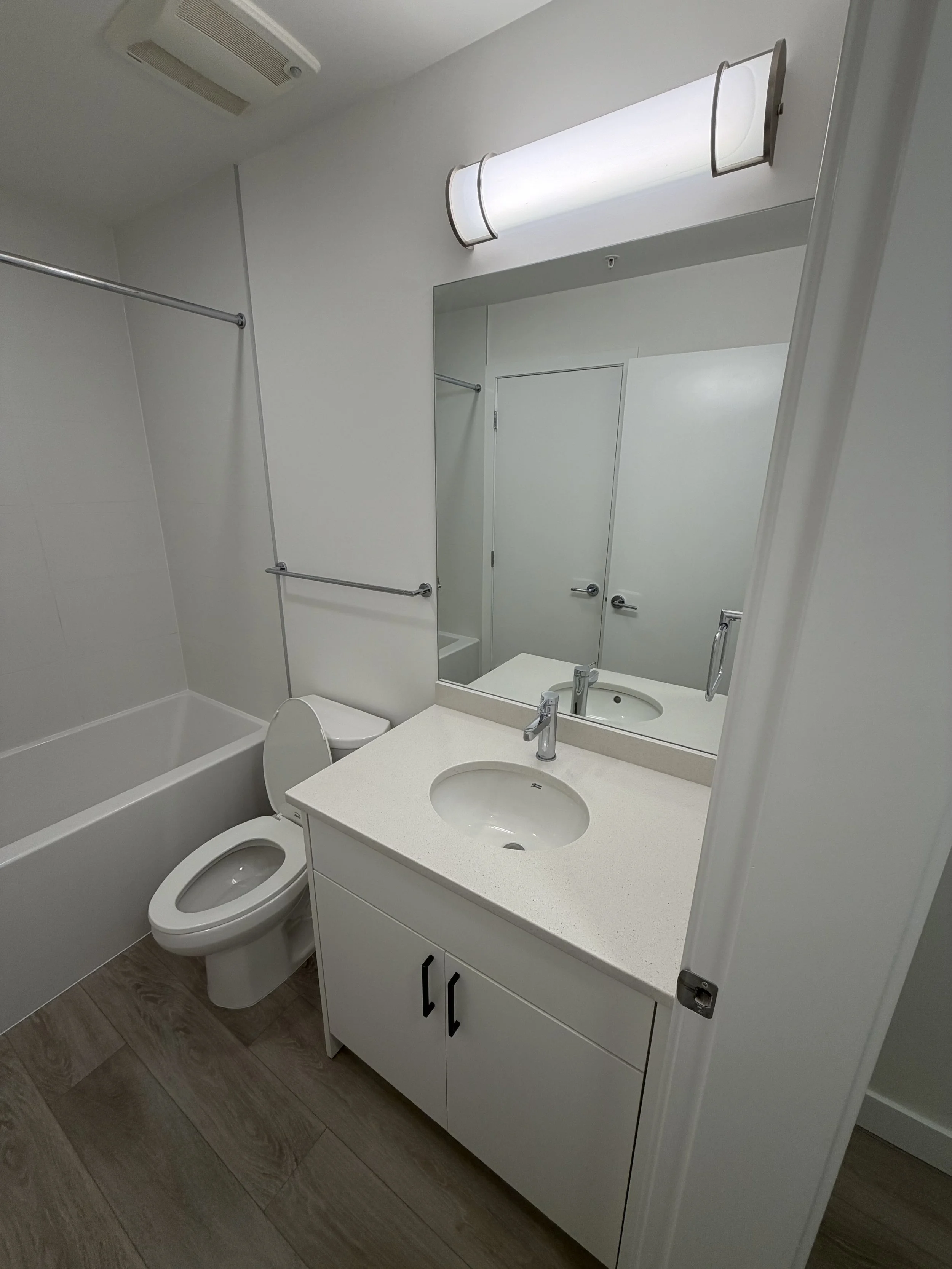 A clean bathroom with a bathtub, toilet, and a white vanity with a sink. A large mirror above the vanity and a frosted light fixture above the mirror. The bathroom has gray flooring and white walls.