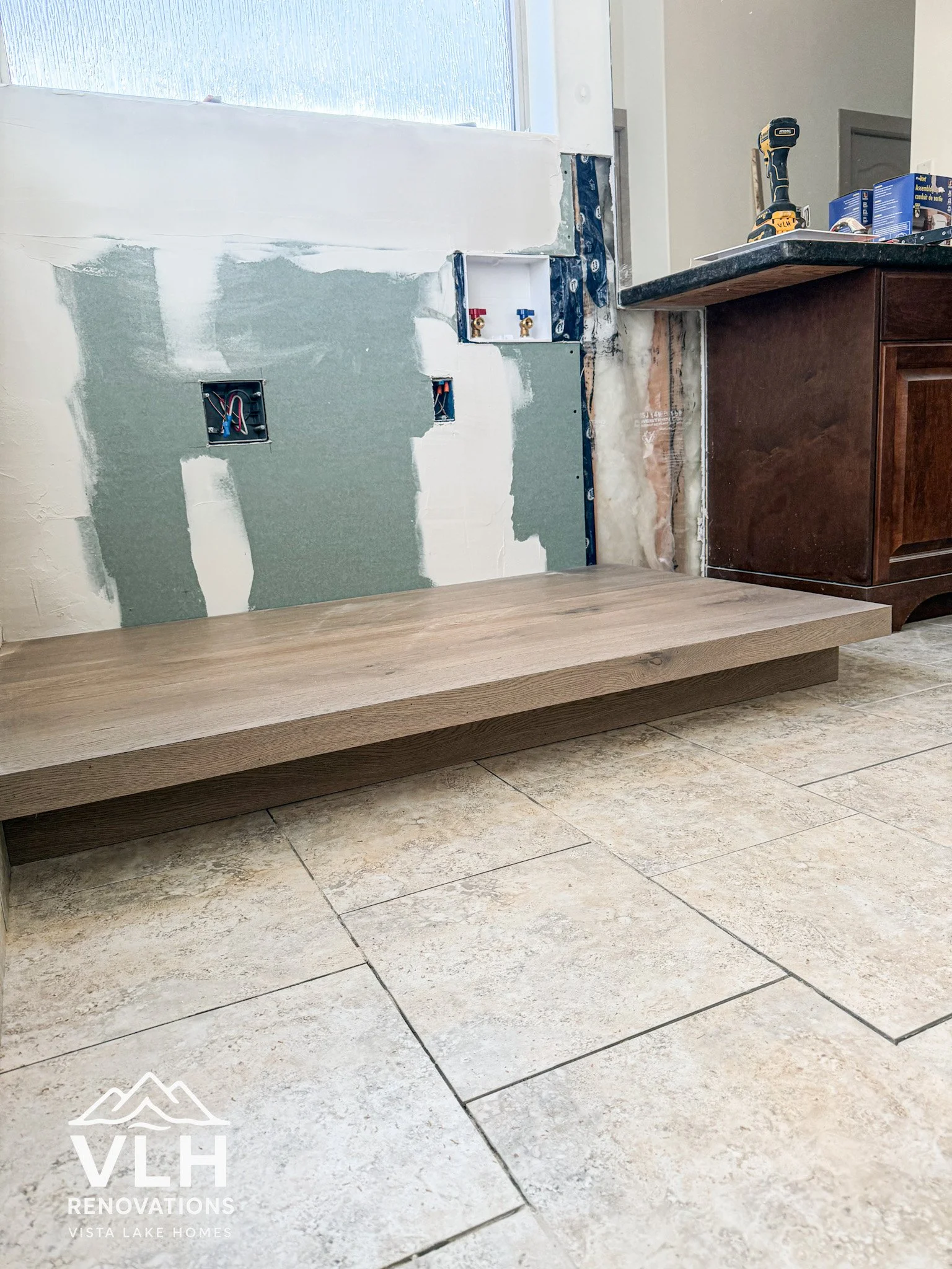 Construction area showing partially installed wooden baseboard, electrical wiring in wall boxes, and ongoing wall repairs. A countertop with a drill and other tools is visible, and the floor is tiled.