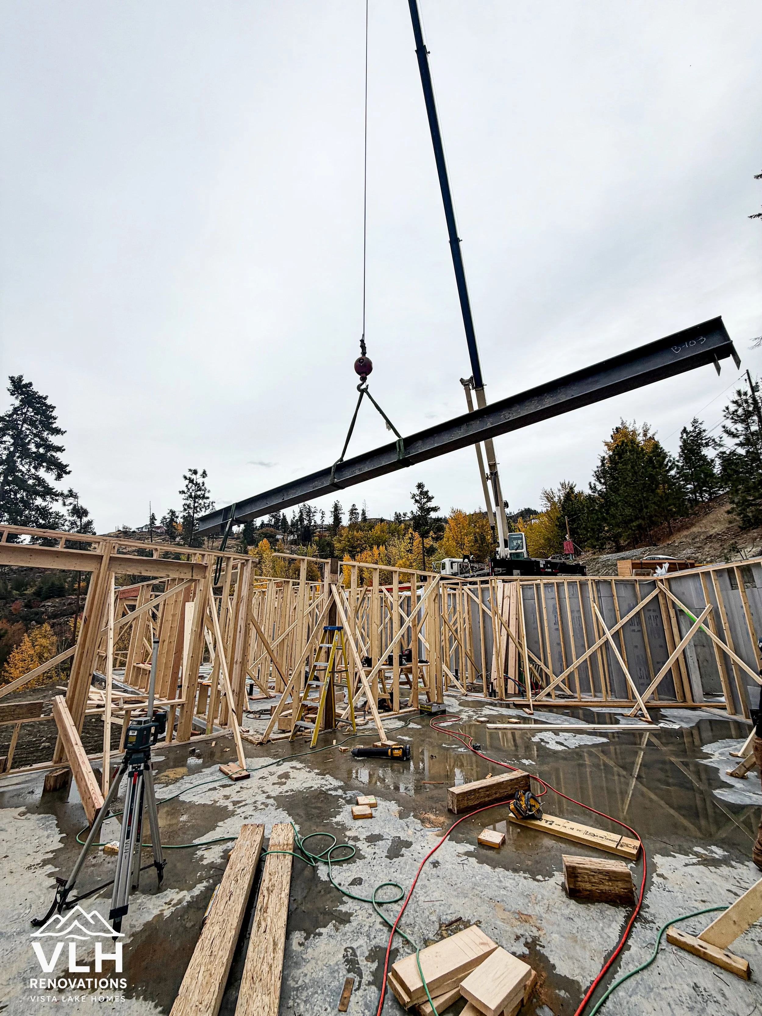Construction site with wooden framing, a crane lifting a steel beam, and construction tools and materials on wet concrete ground.
