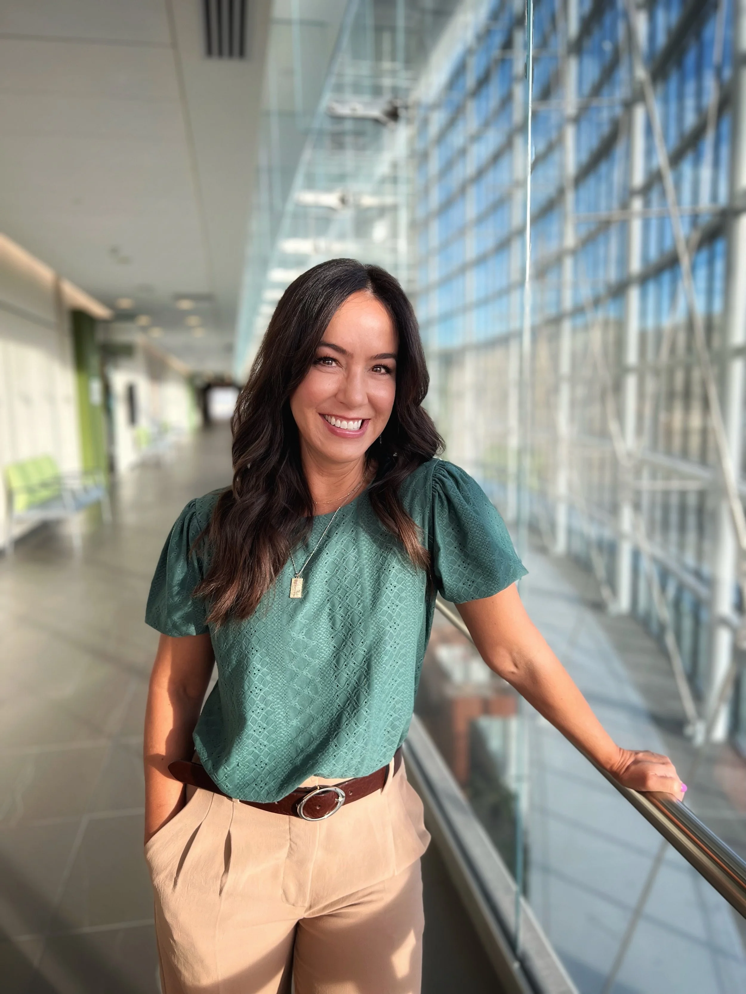 Smiling woman with long dark hair wearing a green textured blouse and beige pants, standing by a window in a modern building.