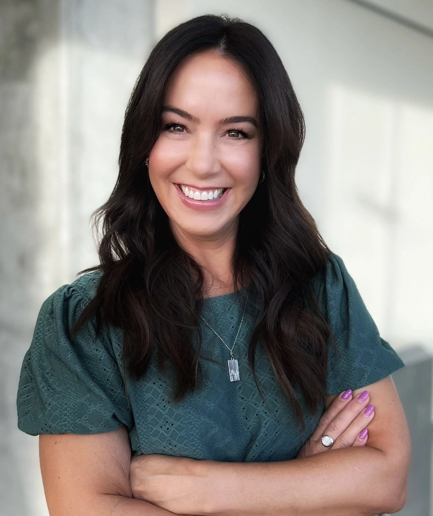 A woman with long dark hair smiling, wearing a teal dress, with arms crossed in front of a bright background.