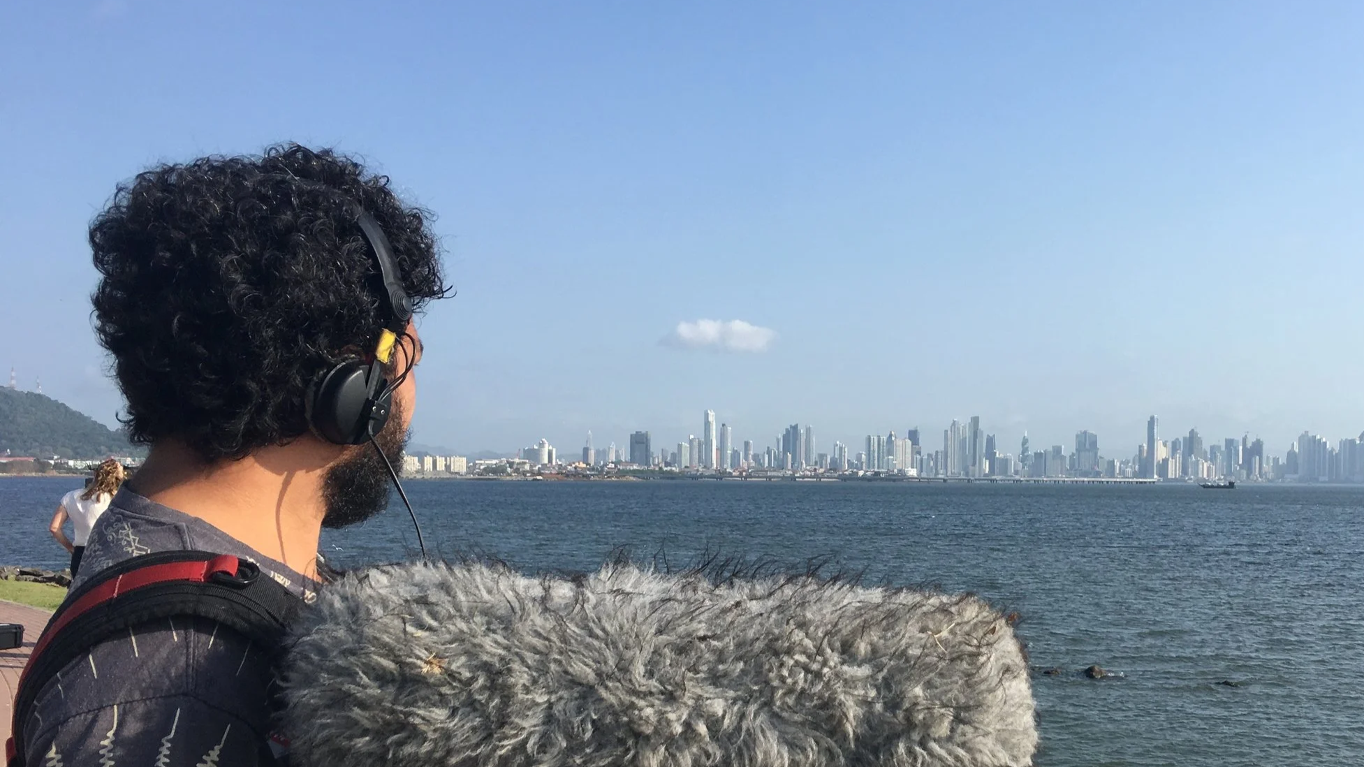 Ingmar Herrera Hombre con auriculares y microfono grabando el. skyline observando el horizonte de la ciudad de Panamá desde un parque junto al agua, con un parque y un cielo despejado en el fondo.