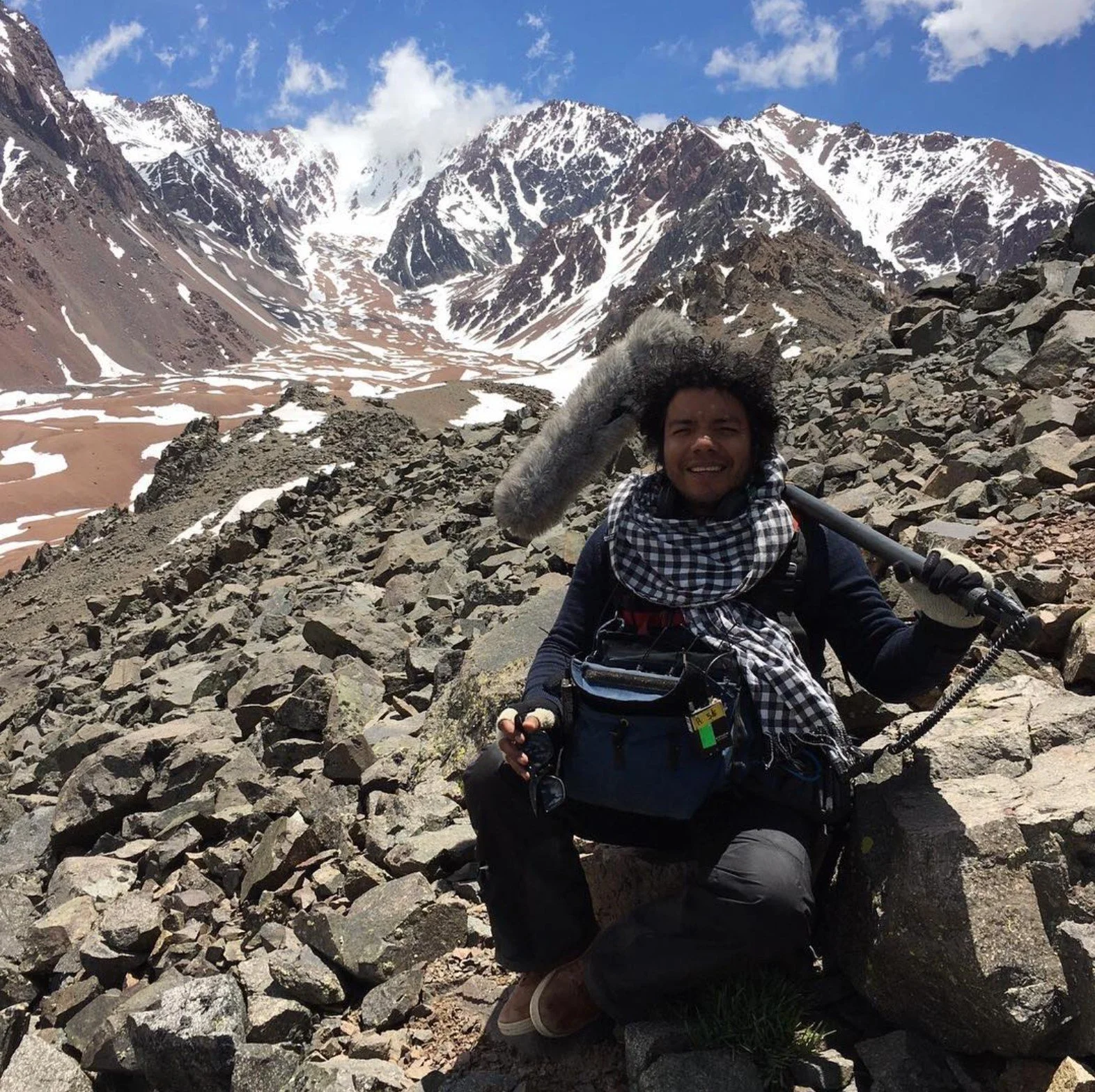 Un hombre con cabello rizado y barba, abrigado con bufanda y guantes, sosteniendo un Micrófono en un paisaje de montañas cubiertas de nieve y rocas, con cielo azul y algunas nubes. Aconcagua Ingmar Herrera Graba sonidos