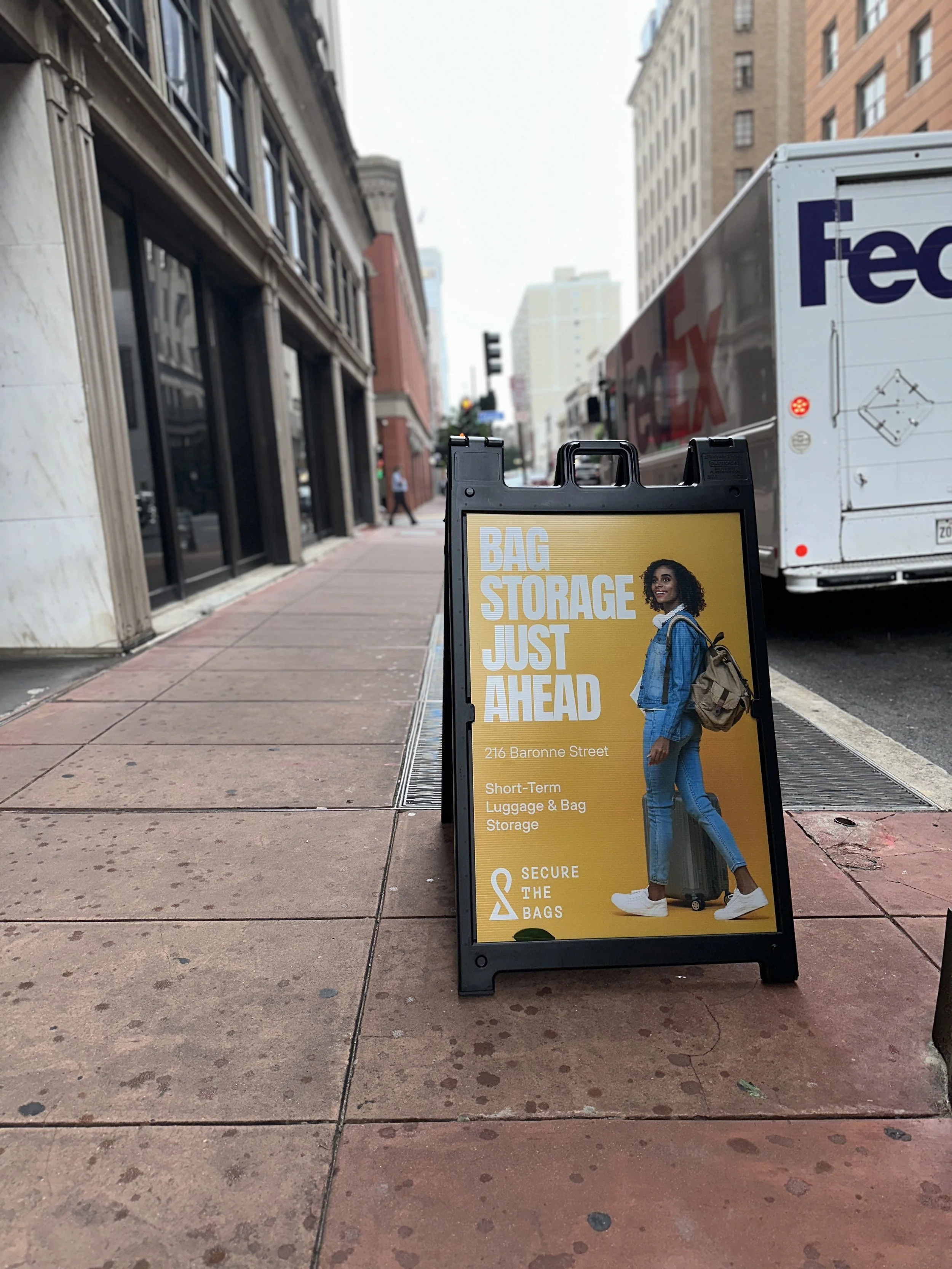 A sidewalk sign advertising short-term luggage and bag storage located at 216 Baronne Street, featuring a smiling woman with curly hair, a blue denim jacket, jeans, white sneakers, and a tan backpack, with a city street and buildings in the background.