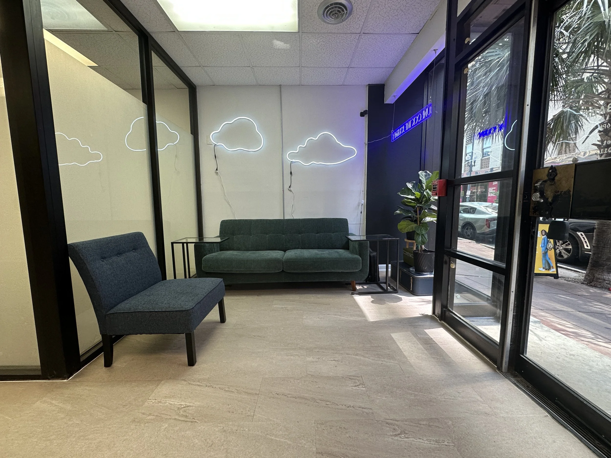 Interior of a modern waiting area with a green sofa, a blue armchair, side tables, a potted plant, neon cloud lights on the wall, and a large glass front door.