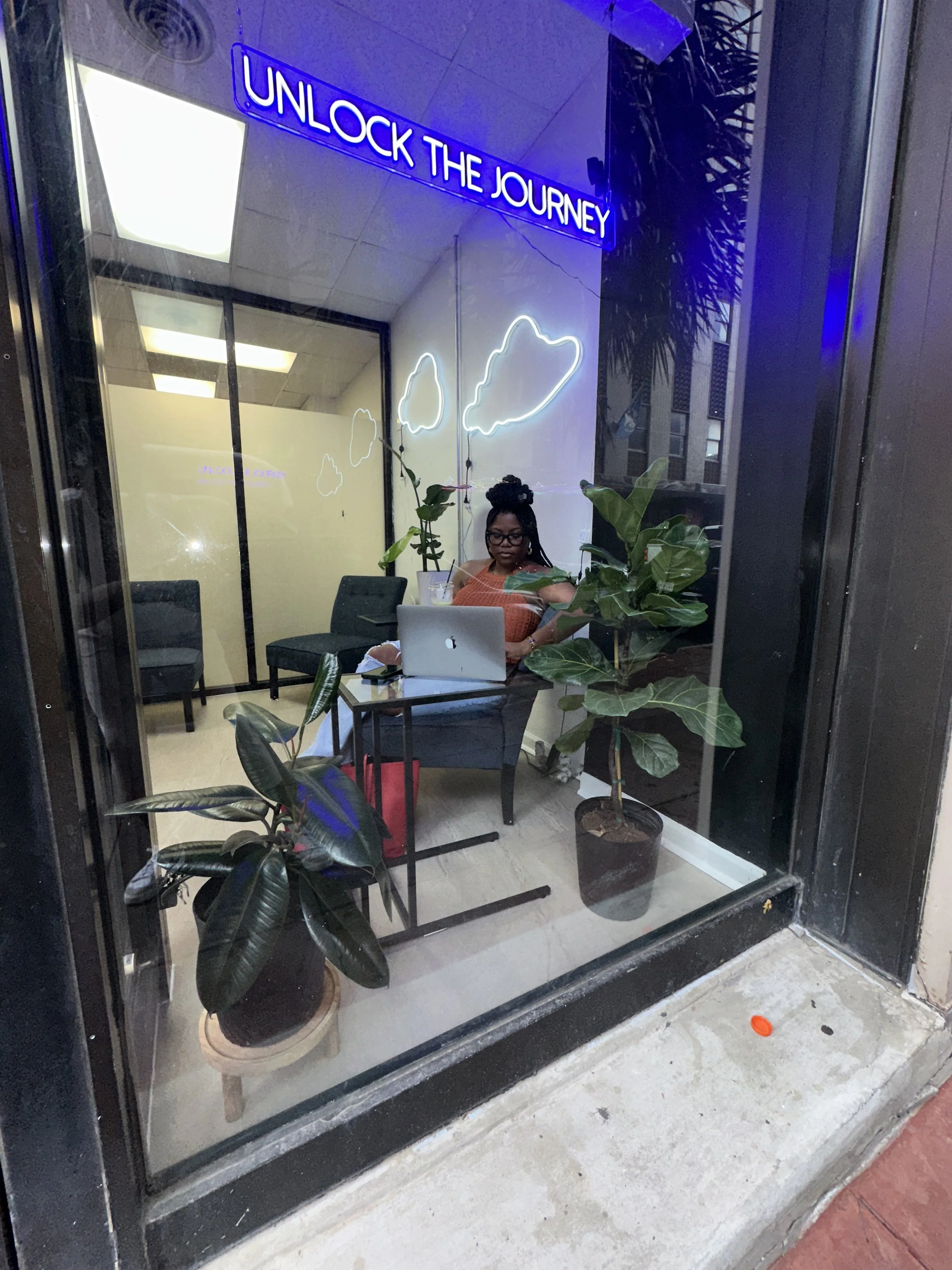 A woman sitting at a desk inside a room with glass walls, working on a laptop. There are plants inside the room and neon signs that say 'UNLOCK THE JOURNEY' and cloud shapes on the wall.