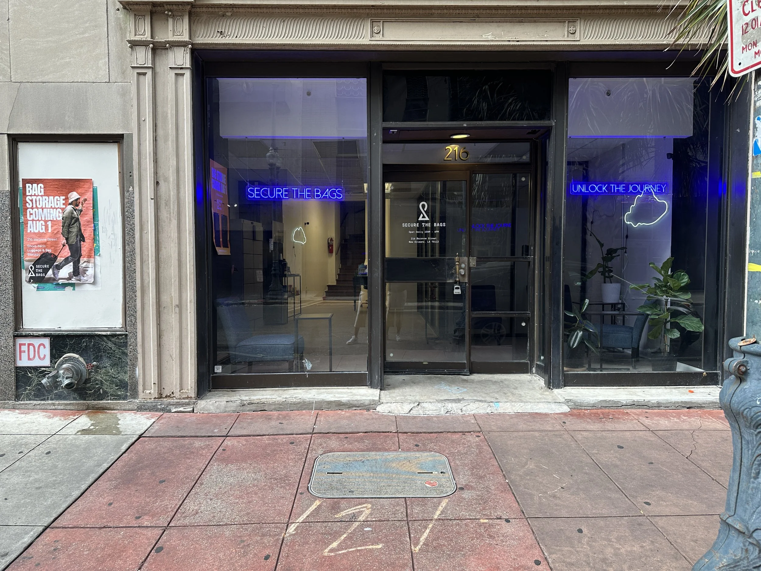 storefront with glass doors and windows, neon signs saying "Secure the Bags" and "Unlock the Journey," potted plants inside, sidewalk with painted lines, poster on the left advertising upcoming bag storage service, and a blue fire hydrant on the right.