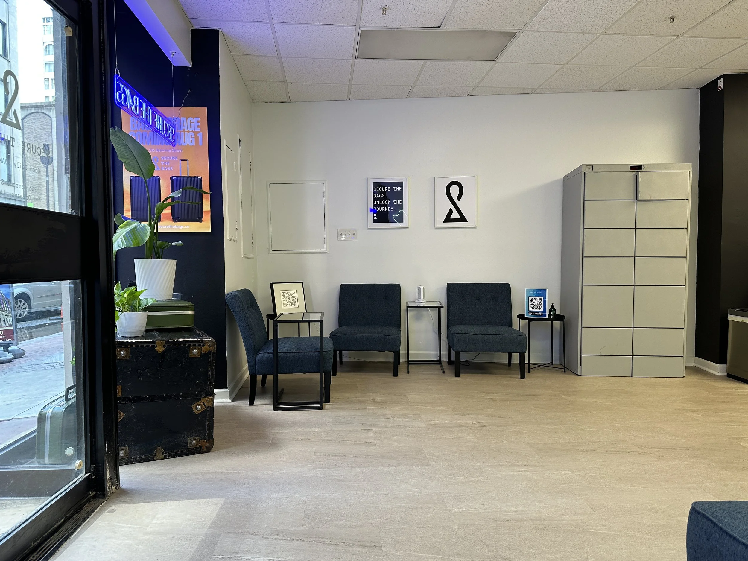 Empty waiting room with three blue chairs, a side table with a QR code, a large storage cabinet, and wall art including a logo and informational posters. A plant and suitcase near the entrance, with a glass door showing the street outside.