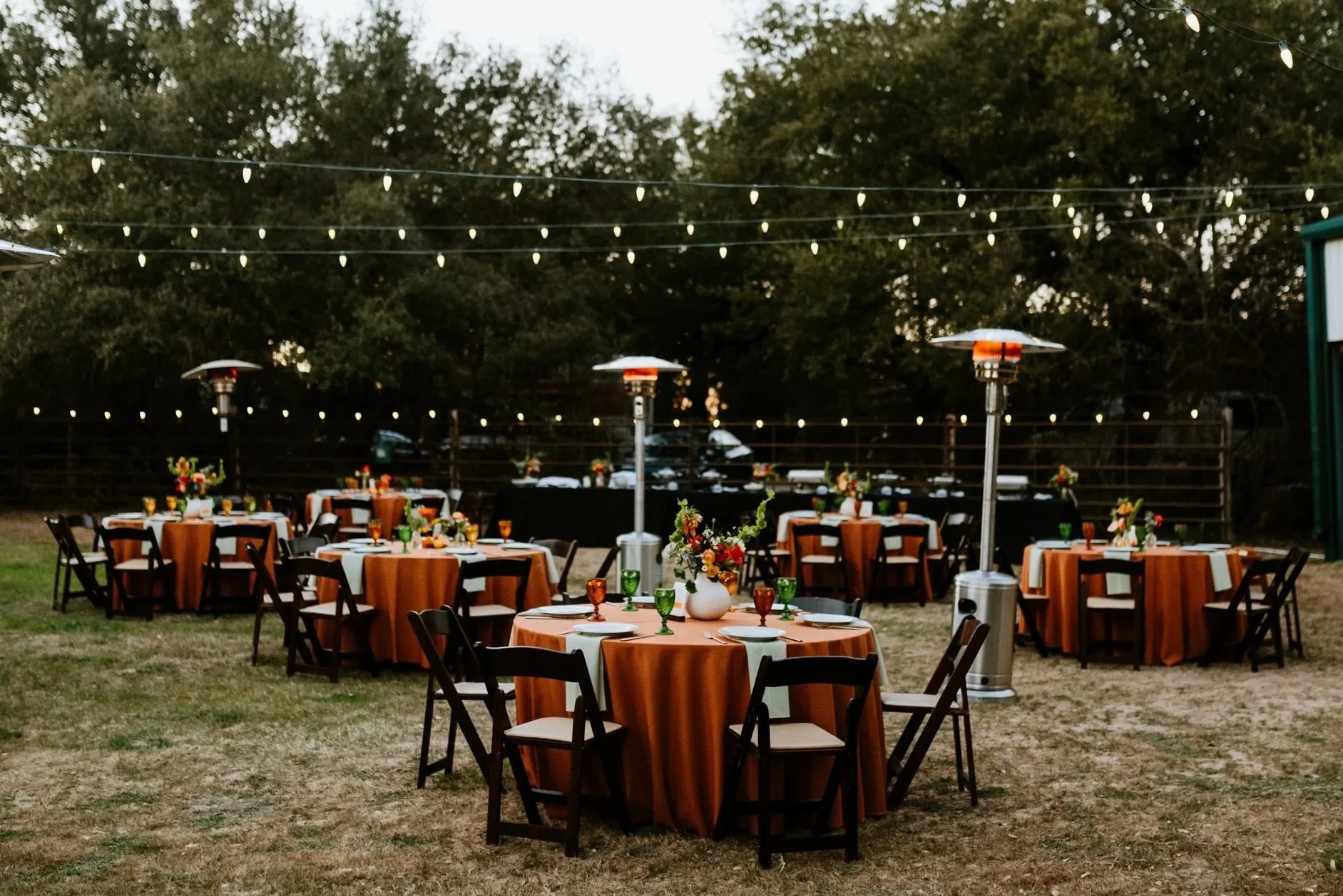 Outdoor event setup with round tables covered in orange tablecloths, surrounded by black chairs, under string lights. Each table has a floral centerpiece and a heater nearby. Trees are visible in the background.