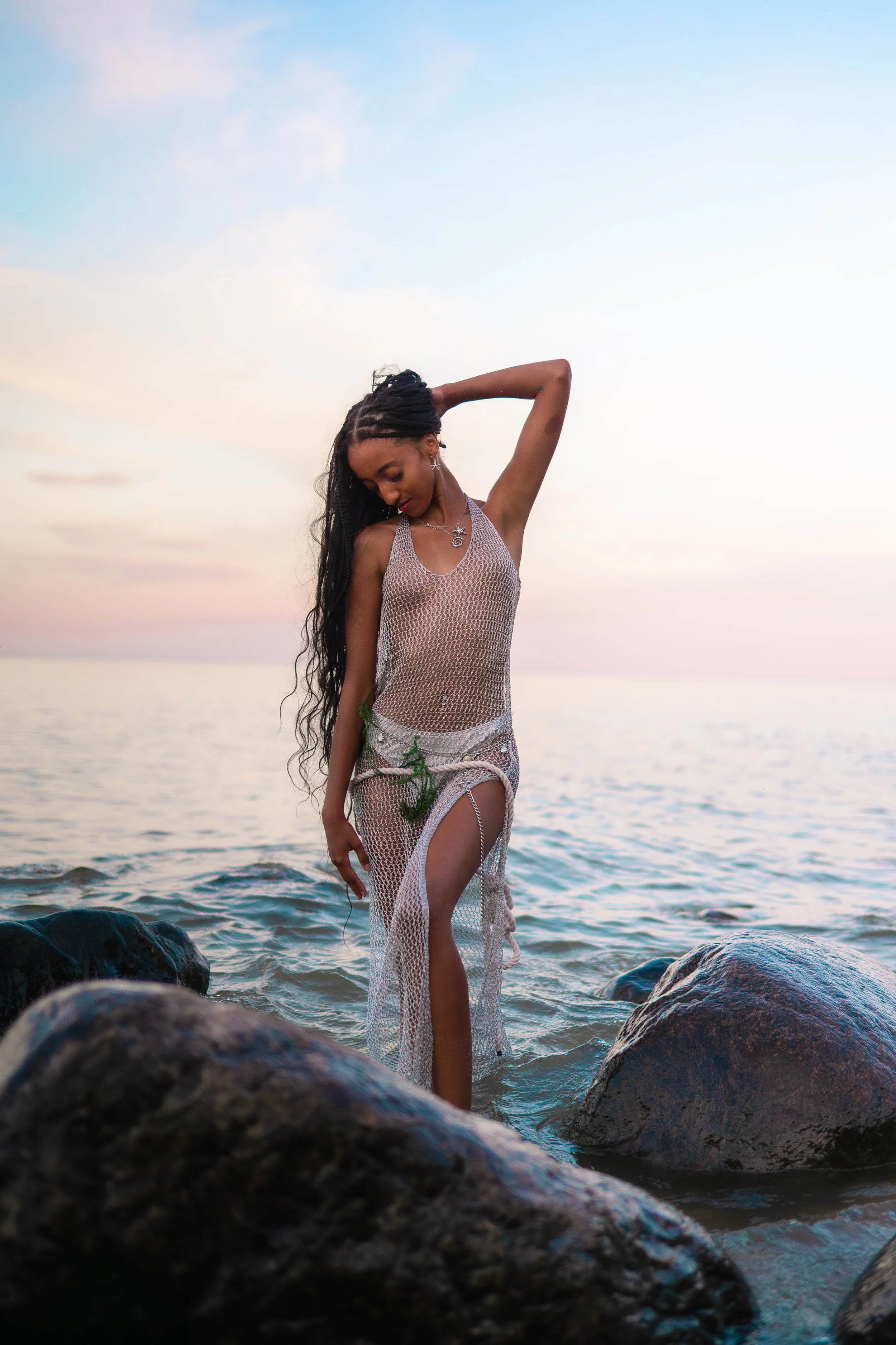 A woman standing on rocks in a body of water at sunset, wearing a see-through crocheted outfit and jewelry, with long, dark hair and a relaxed pose.