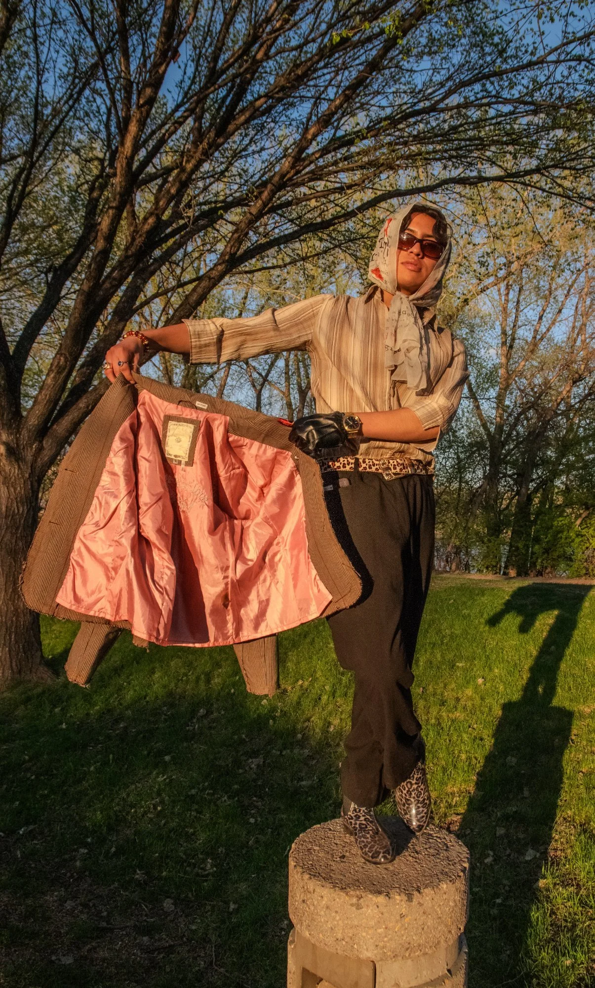 A person with dark hair, wearing sunglasses, a headscarf, striped shirt, black pants, leopard print shoes, and a watch, stands on a concrete block outdoors holding an opened brown jacket with pink lining.