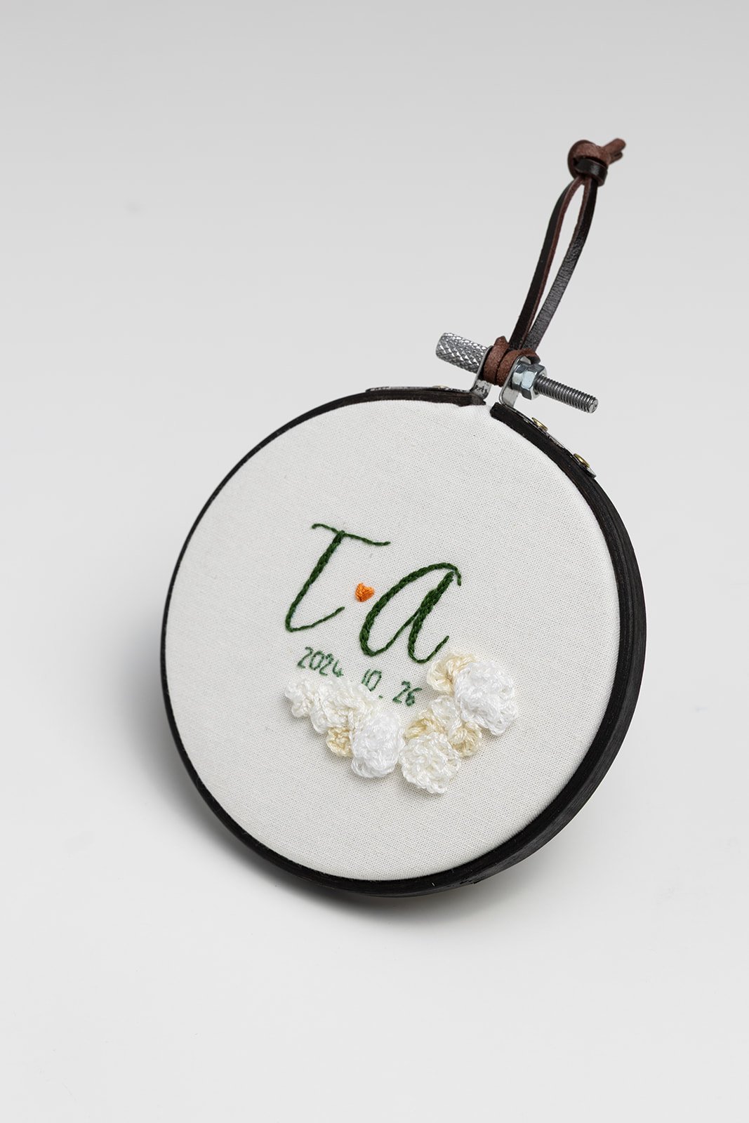 Embroidery hoop with initials 'T' and 'A' stitched in green, the date '2024.10.26', a small heart, and white crocheted flowers on white fabric.