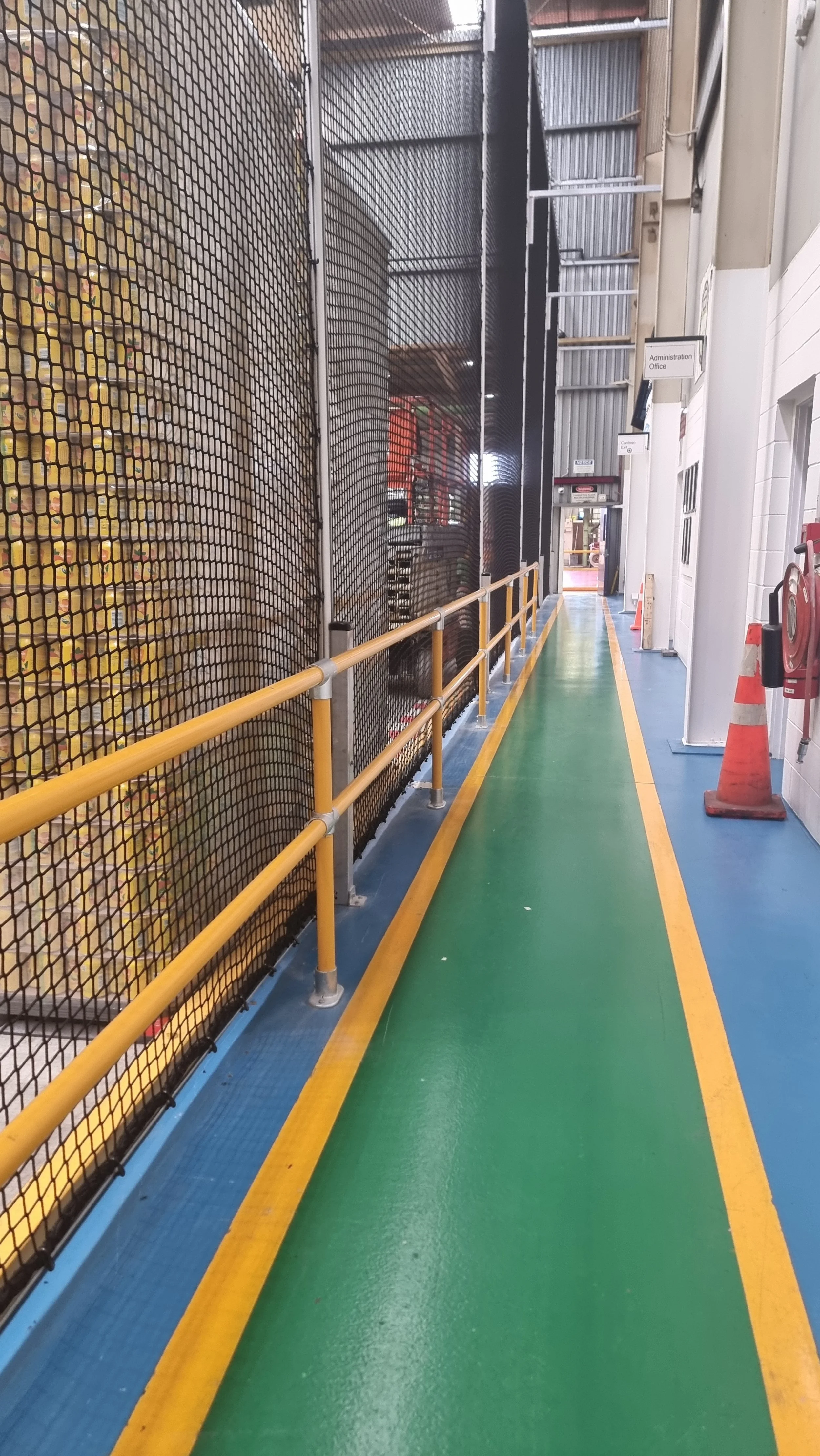 Indoor walkway with green and blue flooring, yellow safety rails, and a black mesh fence on the left side. 